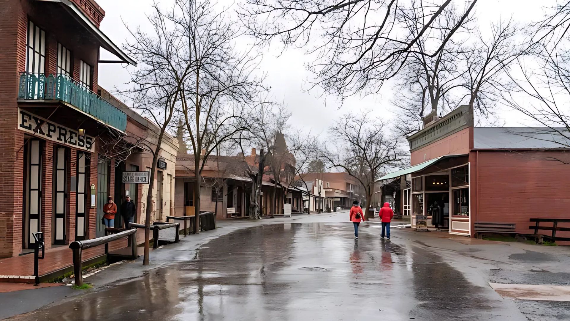 A wide shot of a historic street in Columbia State Historic Park on a wet, overcast day, with two people in red jackets walking away from the viewer down the center of the wet street. On either side, historic brick buildings with wooden boardwalks and bare trees line the street, with one building on the left prominently displaying "EXPRESS".