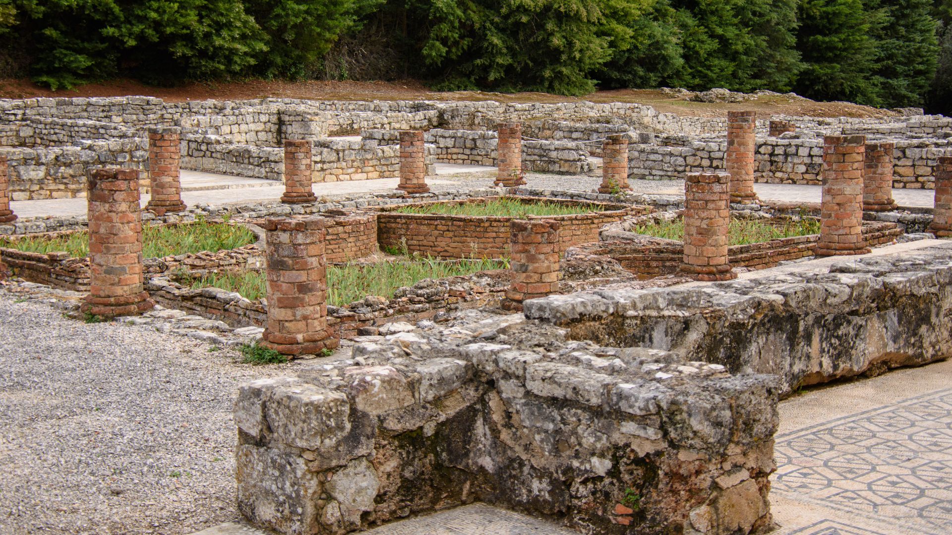 Roman ruins of Conímbriga in Portugal, featuring stone walls, brick pillars, and visible foundations of ancient structures, with some greenery growing within the ruins and trees in the background.