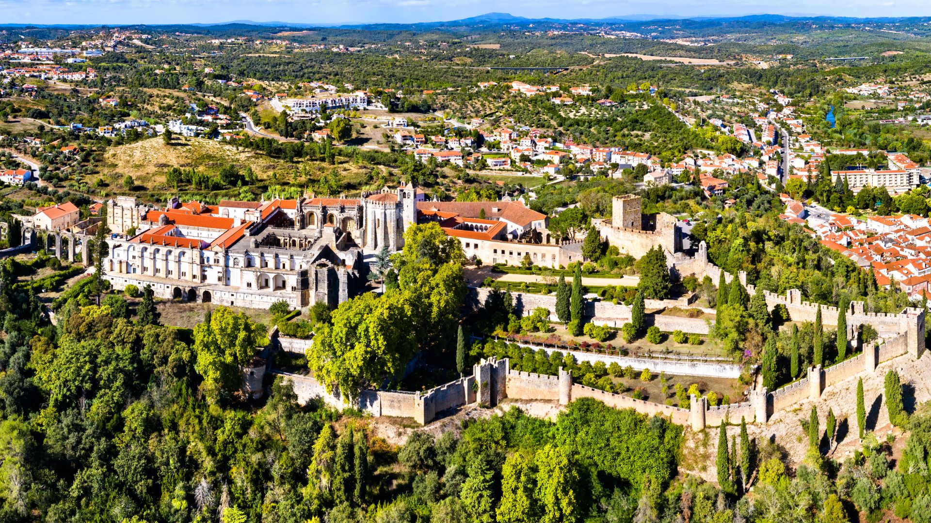 An aerial view showcases the sprawling Convent of Christ and the adjacent Castle of the Knights Templar, a UNESCO World Heritage site, situated atop a hill overlooking the town of Tomar in central Portugal, surrounded by lush greenery and a distant cityscape under a clear sky.An aerial view showcases the sprawling Convent of Christ and the adjacent Castle of the Knights Templar, a UNESCO World Heritage site, situated atop a hill overlooking the town of Tomar in central Portugal, surrounded by lush greenery and a distant cityscape under a clear sky.
