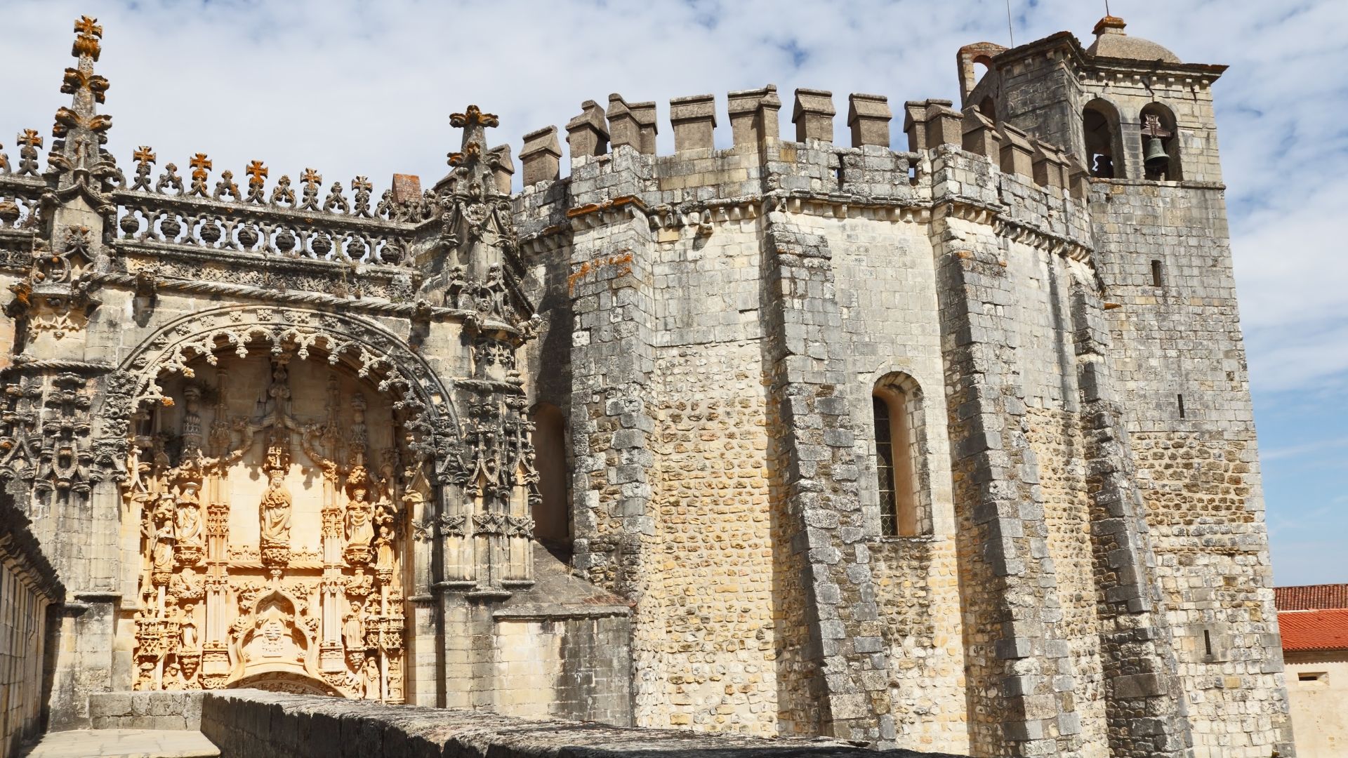 A close-up view of the weathered stone exterior of the Convent of Christ in Tomar, Portugal, showcasing intricate Manueline architectural details on the left and a sturdy, battlemented tower on the right with a bell visible in its belfry.