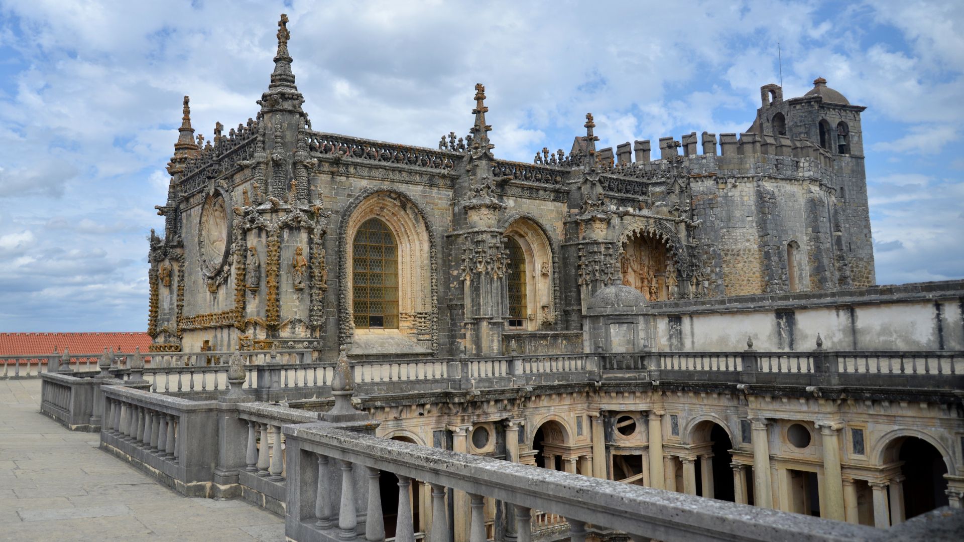 A panoramic view of the Convent of Christ in Tomar, Portugal, showcasing its intricate Gothic and Manueline architecture under a cloudy sky, with a stone balustrade in the foreground.