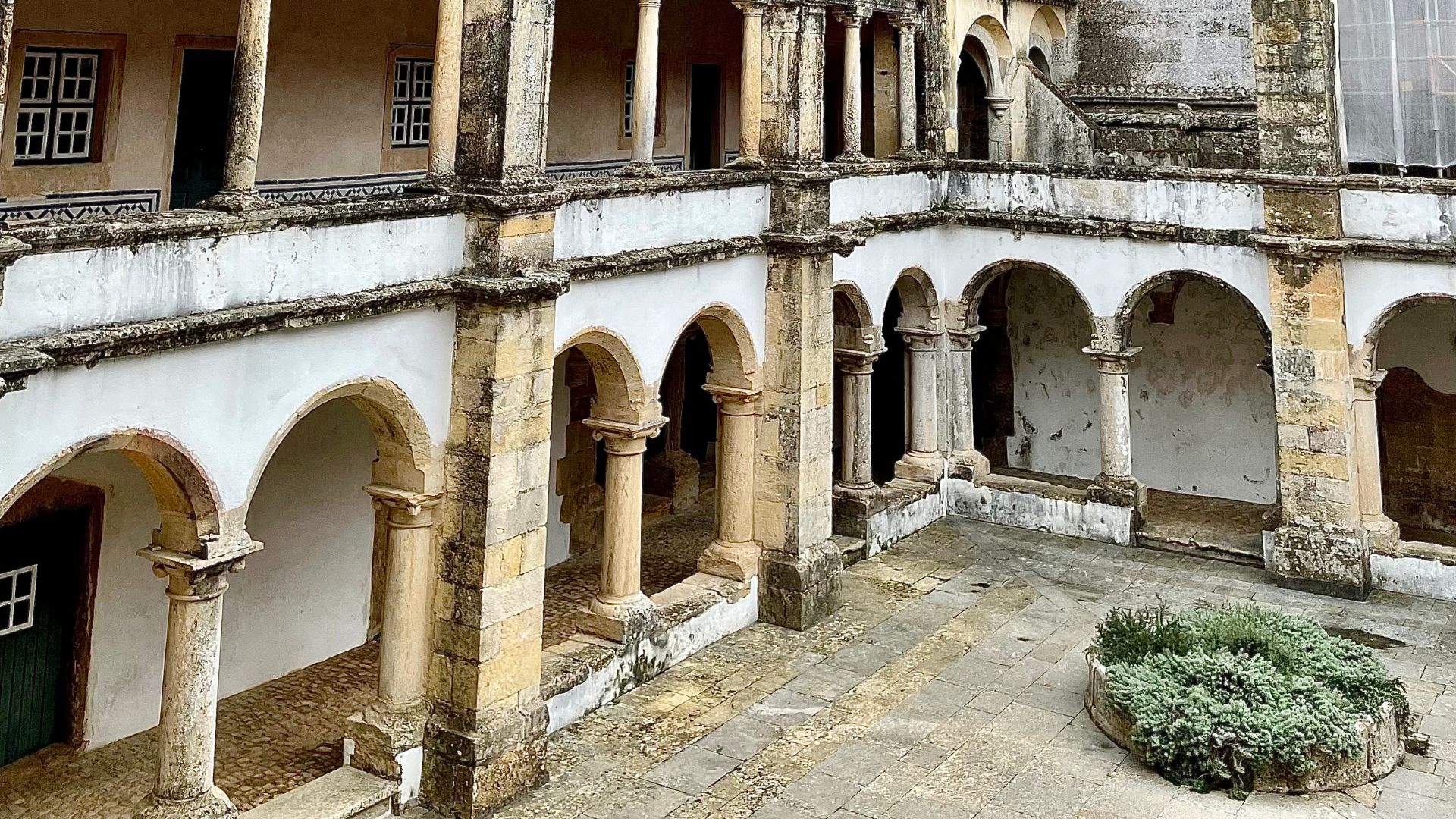 A view of a historic stone courtyard with arched walkways and columns, part of the Convent of Christ in Tomar, Portugal, featuring a small, circular garden bed in the center.
