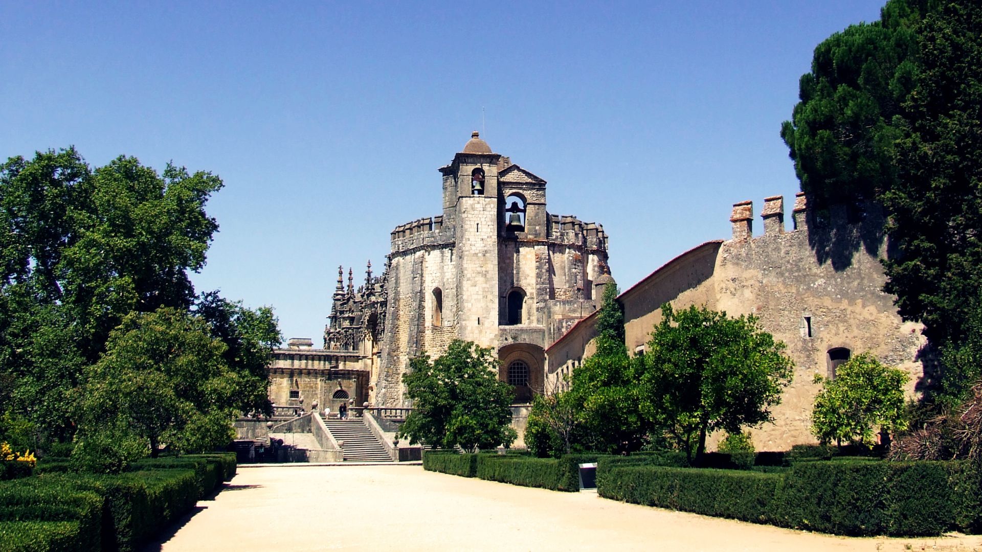 A sunny outdoor shot of the historic Convent of Christ in Tomar, Portugal, showcasing its ancient stone architecture and bell tower, surrounded by green trees and manicured hedges.
