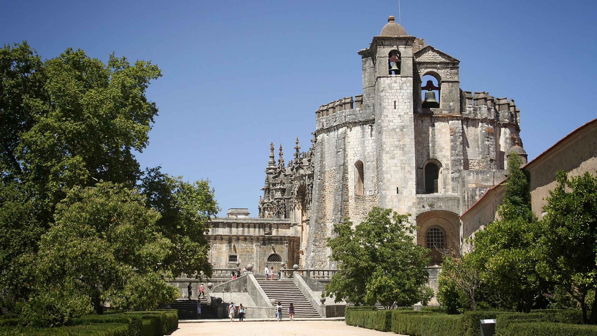A panoramic view of the historic Convent of Christ in Tomar, Portugal, featuring its stone walls, bell tower, and surrounding gardens under a bright sky.