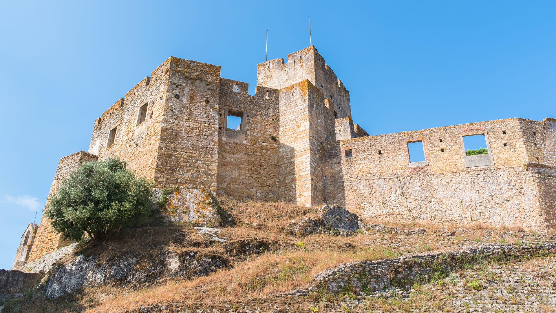 A low-angle view of the ancient, stone-built Convent of Christ or Tomar Castle, a historic complex with weathered walls and window openings, situated on a dry, rocky hillside under a clear blue sky in central Portugal.