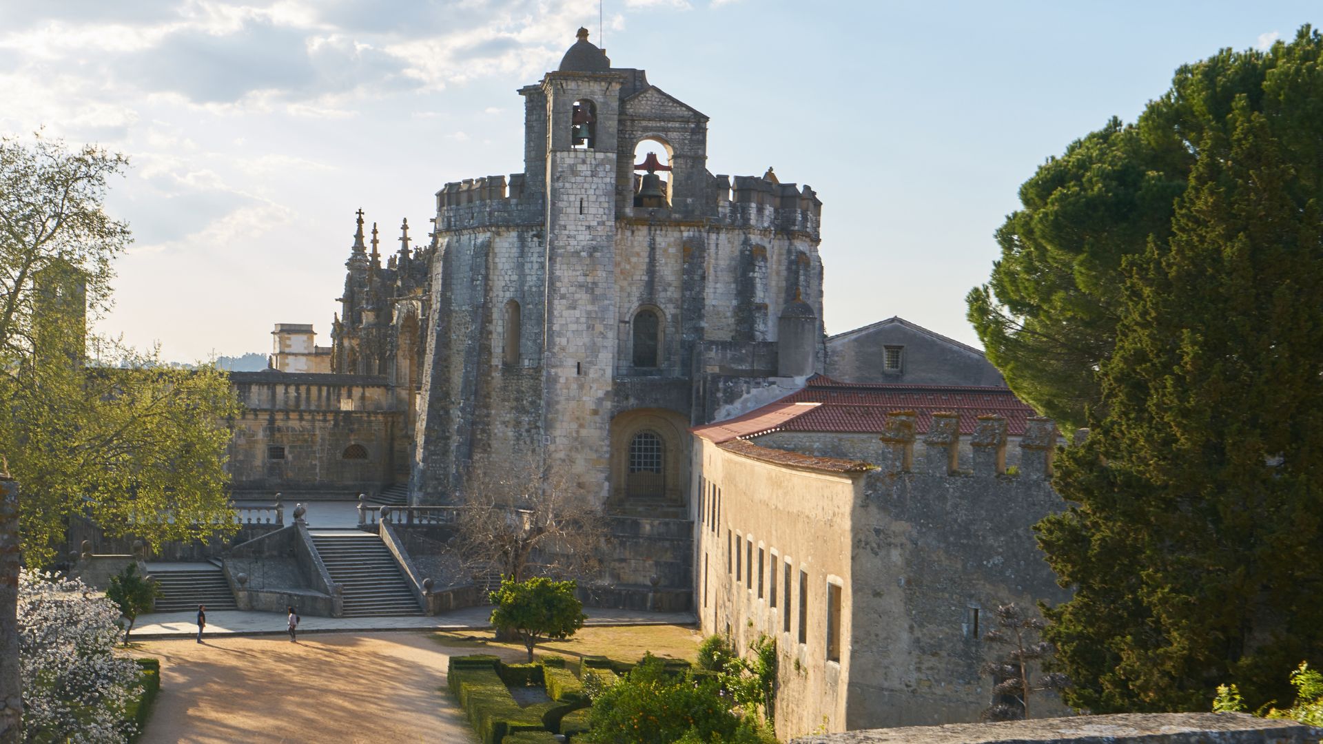A panoramic view of the historic Convent of Christ in Tomar, Portugal, featuring its stone walls, bell tower, and surrounding gardens under a bright sky.