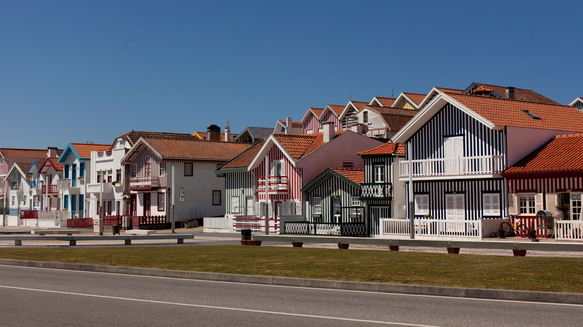 A row of colorful, vertically striped houses with red-tiled roofs line a street in Costa Nova, Portugal, under a clear blue sky.