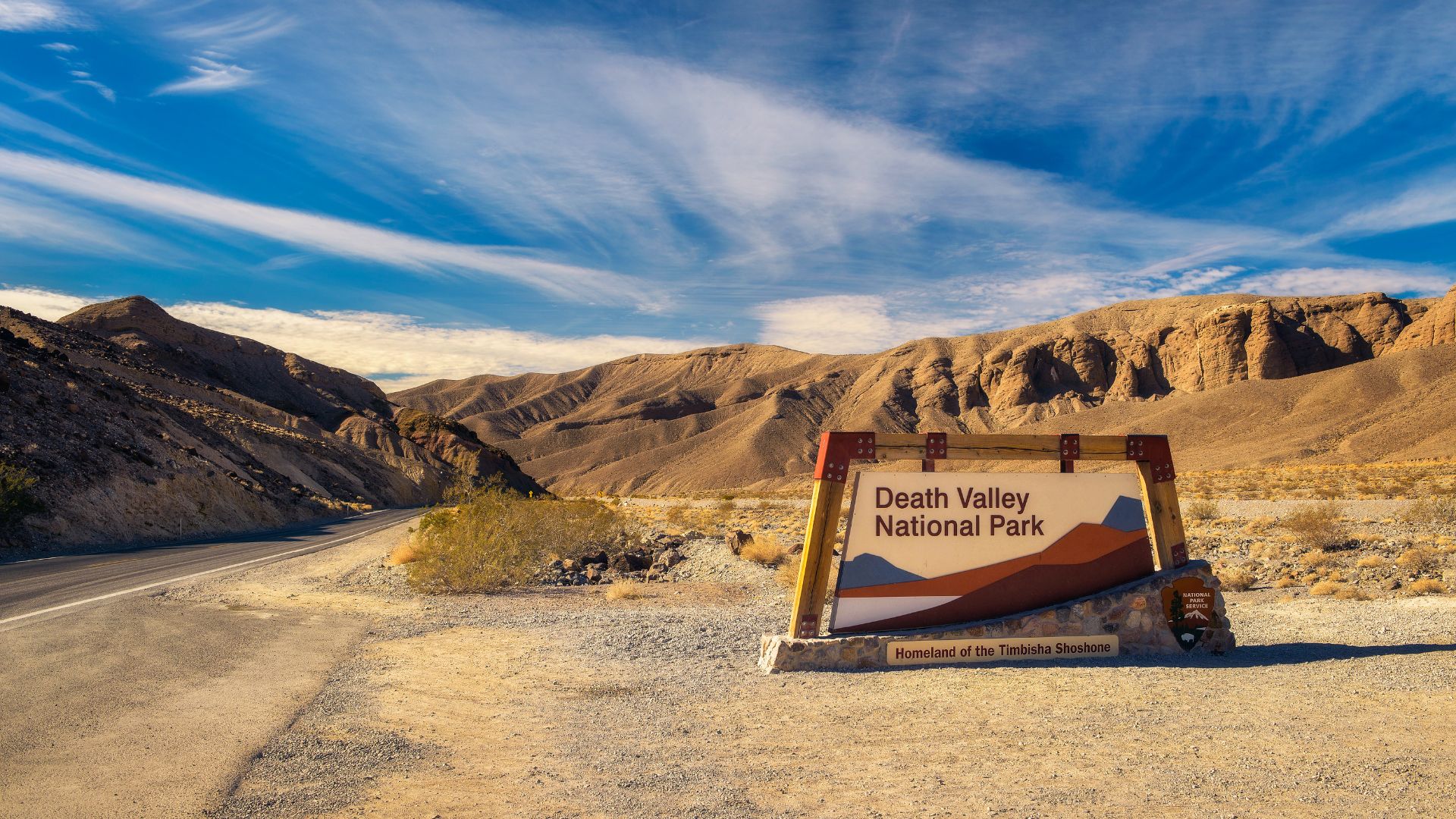 A wide-angle shot of the Death Valley National Park entrance sign in a desert landscape under a blue sky with wispy clouds. The sign, made of wood and rock, features the park's name and an abstract graphic of mountains and a valley. A paved road stretches into the distance on the left, leading towards a range of rugged, barren mountains under a bright sky. The foreground is dry, sandy terrain with sparse desert vegetation.