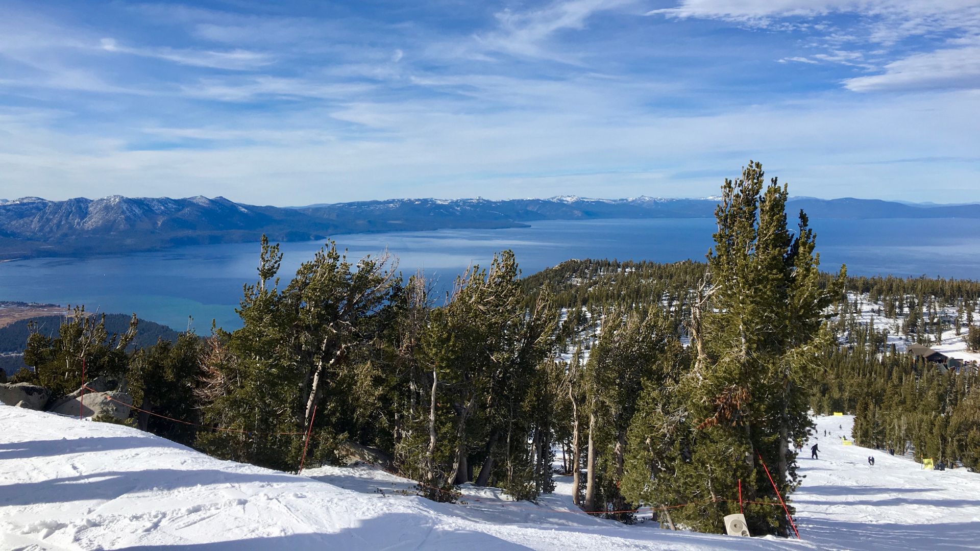 A snowy mountain slope with evergreen trees in the foreground, overlooking the vast blue waters of Lake Tahoe under a partly cloudy sky, with distant mountains visible across the lake.