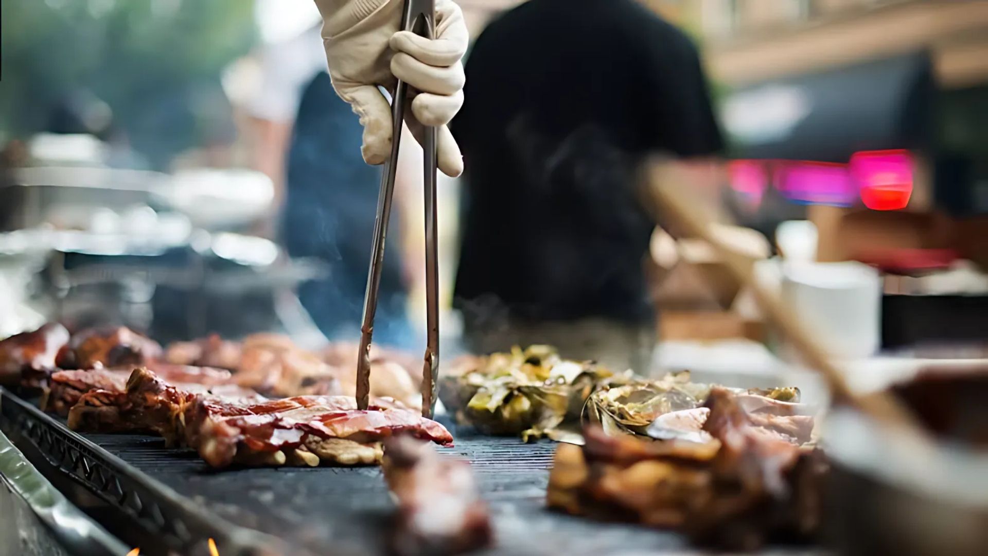 A close-up shot of a person in white gloves using tongs to turn ribs and chicken on a large outdoor grill, with other food items and a blurred background of people and lights.