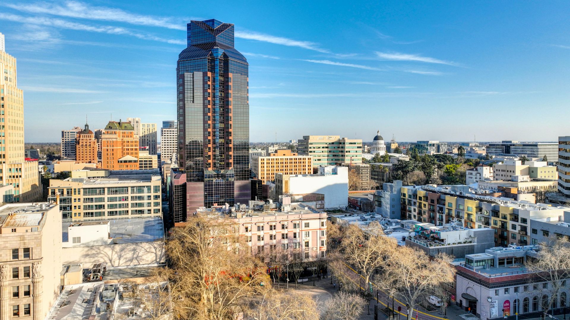A high-angle view of the downtown Sacramento skyline under a clear blue sky, featuring the tall, dark-glassed Renaissance Tower (also known as 500 J Street) in the center, surrounded by other multi-story buildings, including the historic Citizen Hotel building with its distinctive peak and brick and tile exterior. Bare trees are visible in the foreground, indicating winter, and the California State Capitol dome can be seen in the distance on the right.