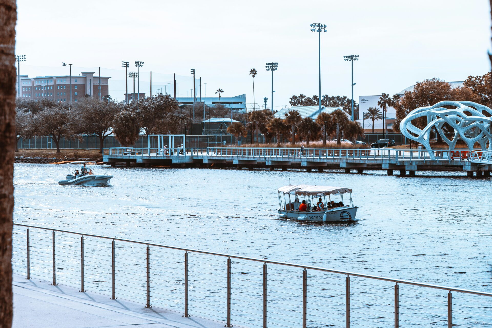 Serene view of Tampa Riverwalk at dusk, with a small boat gently gliding along the water under a canopy of soft, twinkling lights. The vibrant city skyline reflects on the river’s surface, creating a romantic and peaceful setting, ideal for a cozy date night.