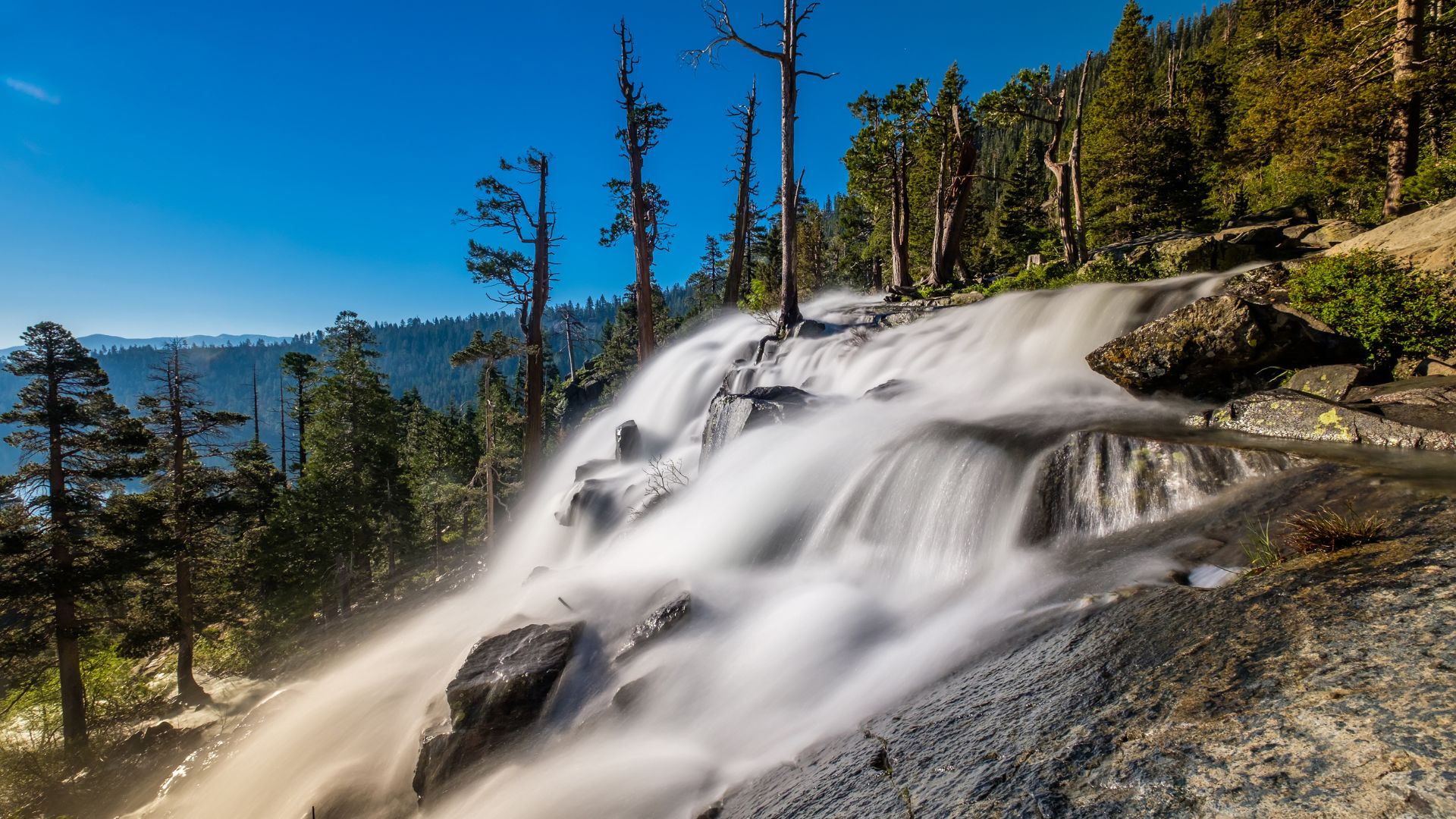 A long-exposure photograph captures the smooth, cascading waters of Lower Eagle Falls flowing over rocks amidst a forested landscape under a clear blue sky.