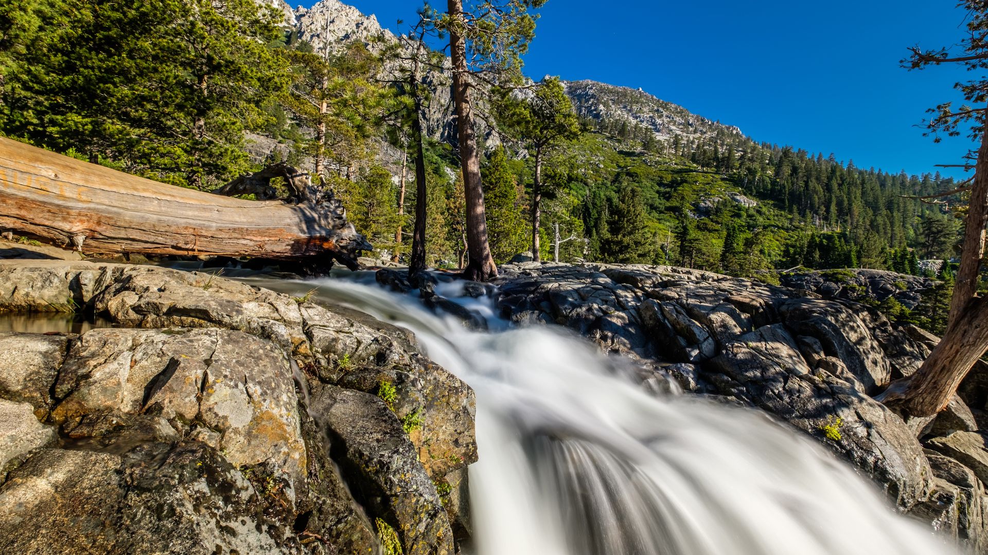 A long exposure photograph of a waterfall cascading over rocky terrain, surrounded by pine trees and mountains under a clear blue sky.