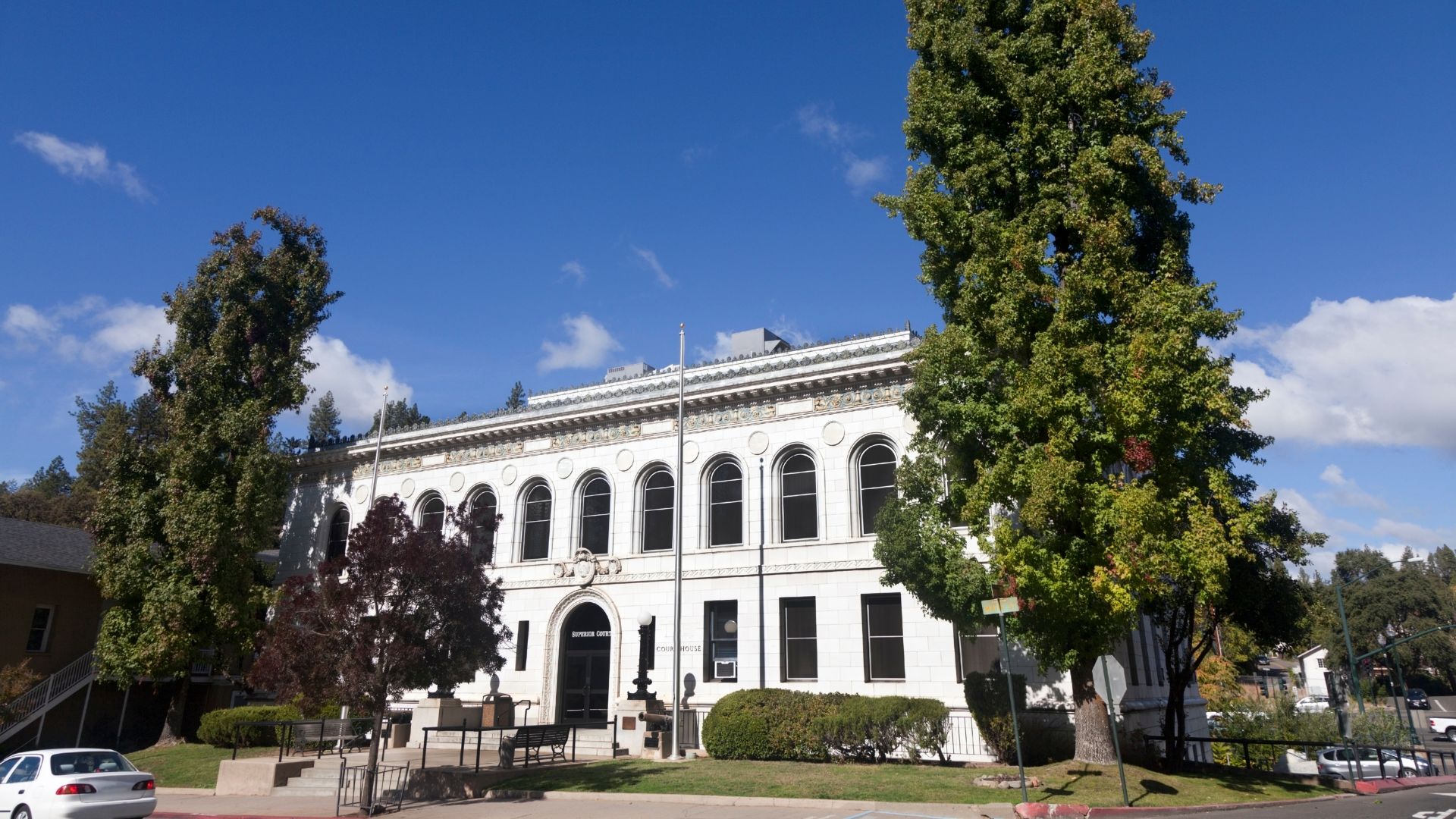 A wide-angle, eye-level shot of the El Dorado County Superior Court building in Placerville, California, a large, white, multi-story building with arched windows and classical architectural details, framed by tall green trees under a clear blue sky with some clouds.