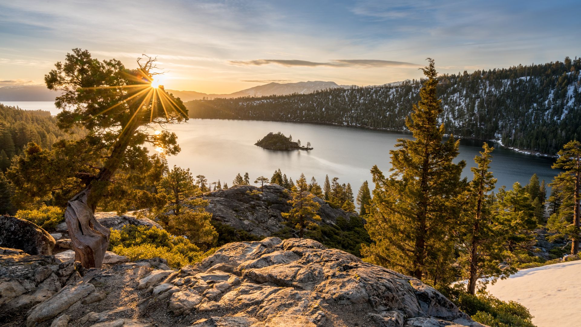 A vibrant sunset casts golden light over Emerald Bay in Lake Tahoe, California, illuminating the calm waters and the tree-covered Fannette Island in the center of the bay. Pine trees and rocky terrain are visible in the foreground, with snow-dusted mountains in the distance under a clear sky.