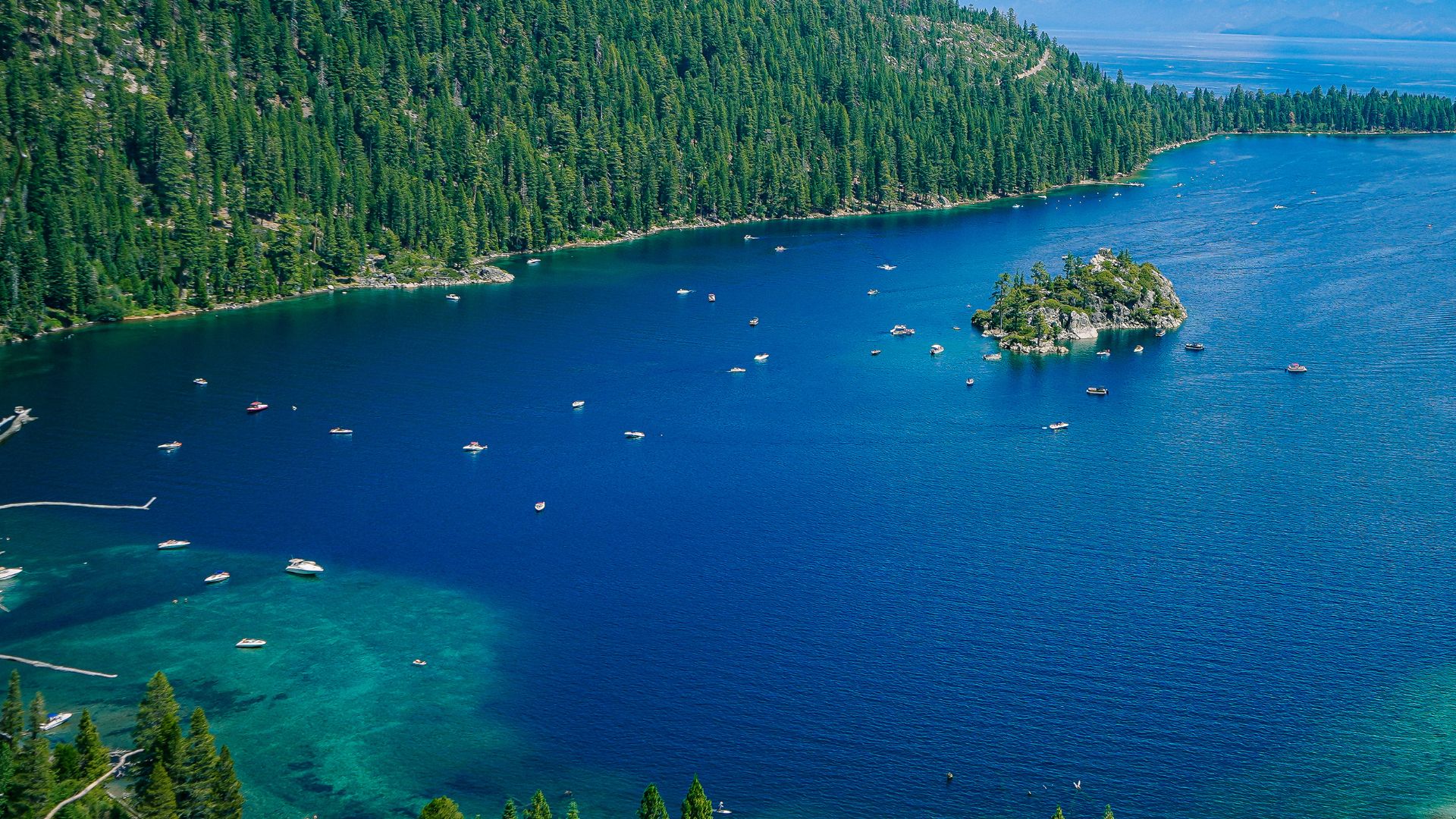 An aerial view showcases the deep blue waters of Emerald Bay at Lake Tahoe, with Fannette Island, a small, tree-covered island, prominently featured in the center. The bay is surrounded by lush green pine forests and mountains, and several boats are scattered across the clear water. 