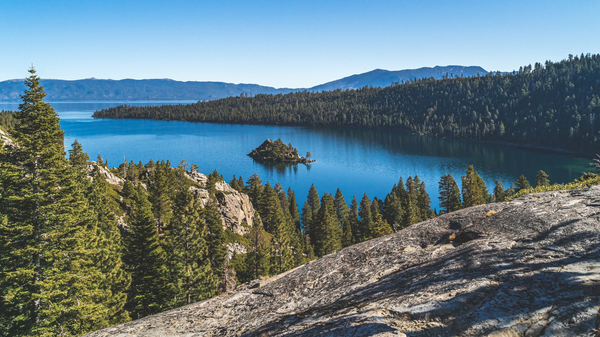 A scenic view of Emerald Bay in Lake Tahoe, showing clear blue water with a small, tree-covered island, surrounded by dense evergreen forests and rocky slopes under a clear blue sky.