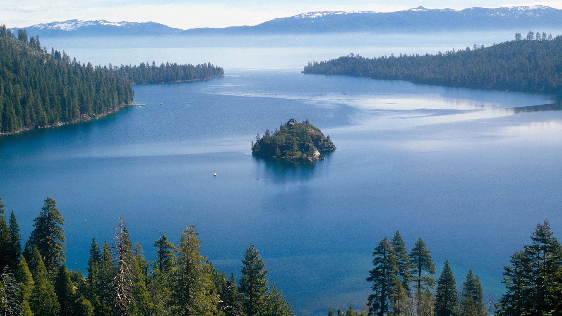 A panoramic view of Emerald Bay in Lake Tahoe, California, showing clear blue water, a small forested island (Fannette Island) in the center, and dense pine forests on the surrounding mountains under a blue sky.