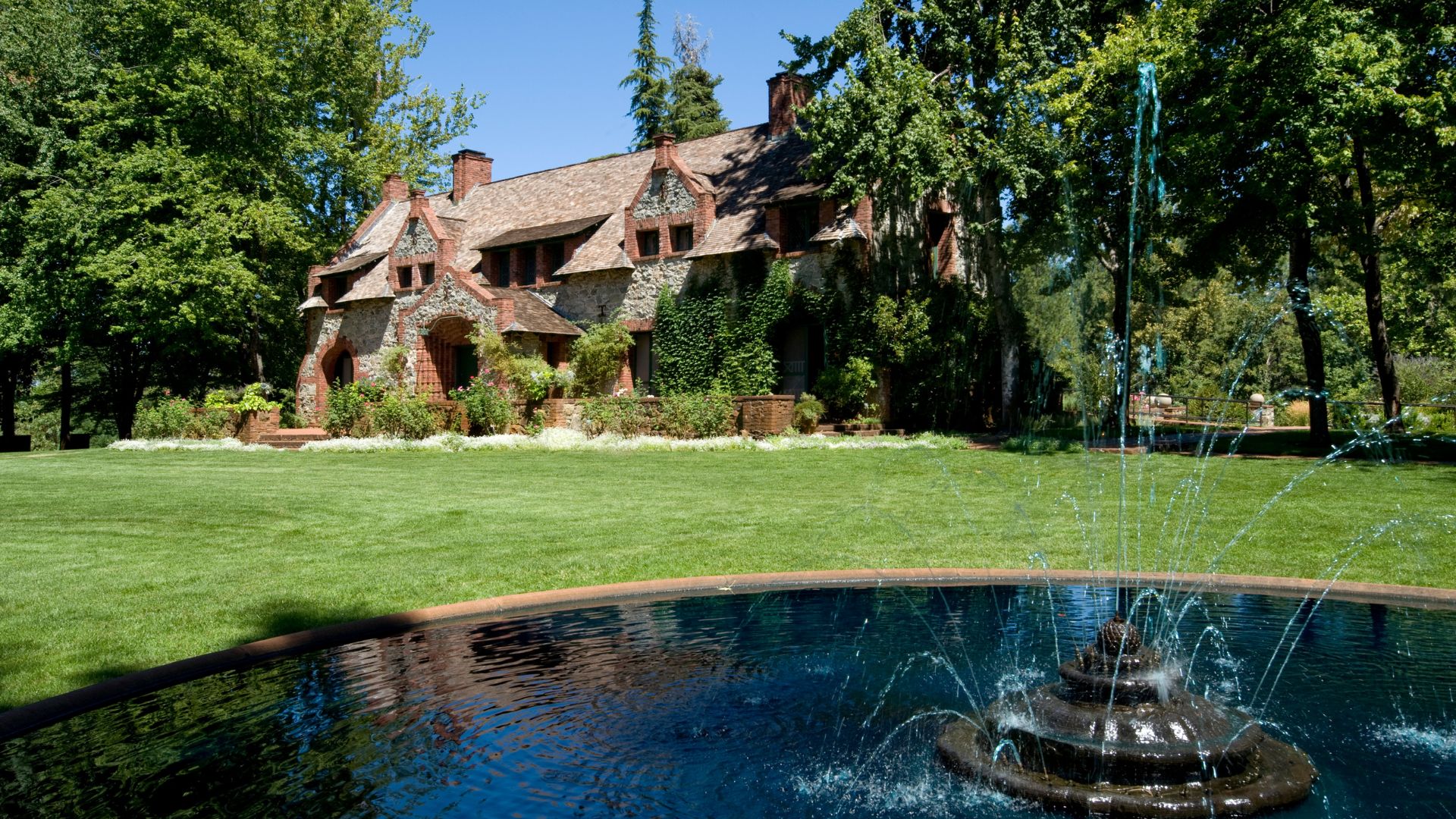 A grand stone house with a gabled roof and multiple chimneys, surrounded by lush green lawns and tall trees, with a tiered fountain in the foreground reflecting the blue sky.