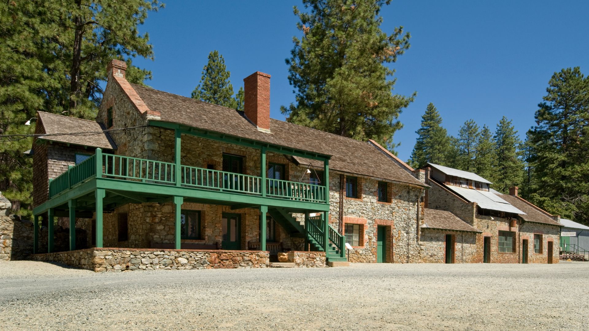 A wide, eye-level shot shows a historic stone building with a green-painted wooden balcony and porch wrapping around the front and side, set against a backdrop of tall pine trees and a clear blue sky. The building features multiple windows, a prominent brick chimney, and a brown shingled roof, with an attached, simpler stone structure extending to the right. The foreground is a flat, light-colored gravel area.