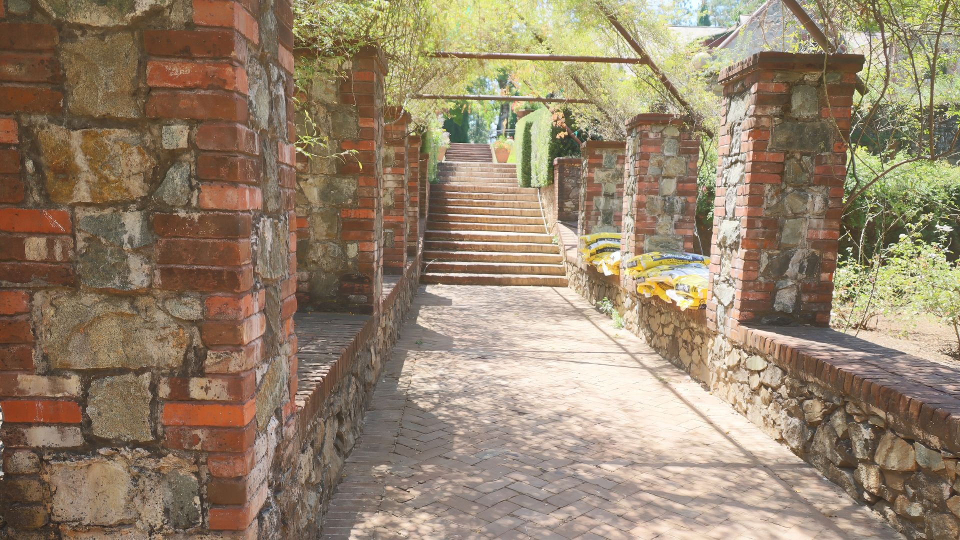A sunny outdoor pathway at Empire Mine State Historic Park, flanked by weathered brick and stone walls with overhanging greenery, leading up a set of stone stairs towards a trellised archway.