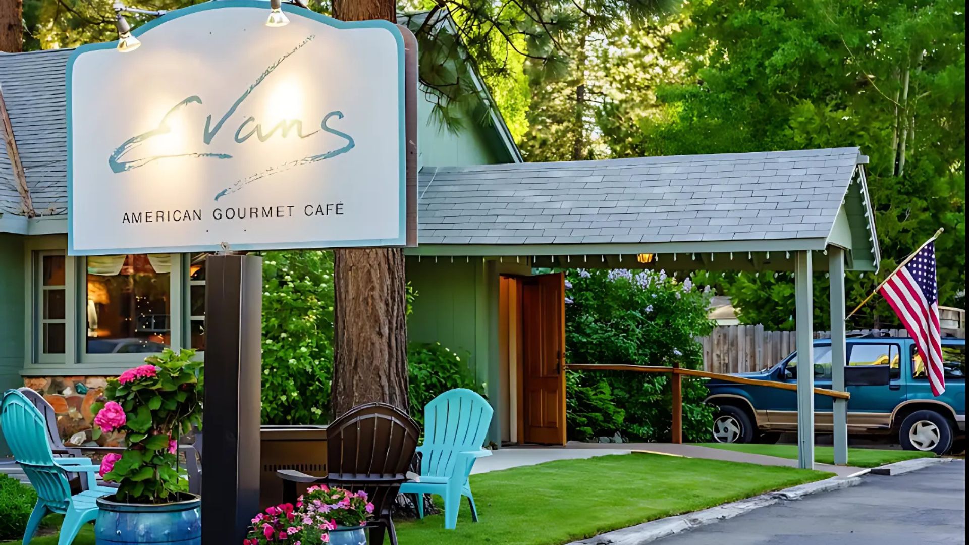 A well-lit sign for "Evan's American Gourmet Cafe" stands in front of a green building with a grey roof, nestled among trees. In the foreground, there are outdoor seating arrangements with light blue chairs and potted plants with pink flowers. An American flag is visible to the right, near a parked blue SUV.