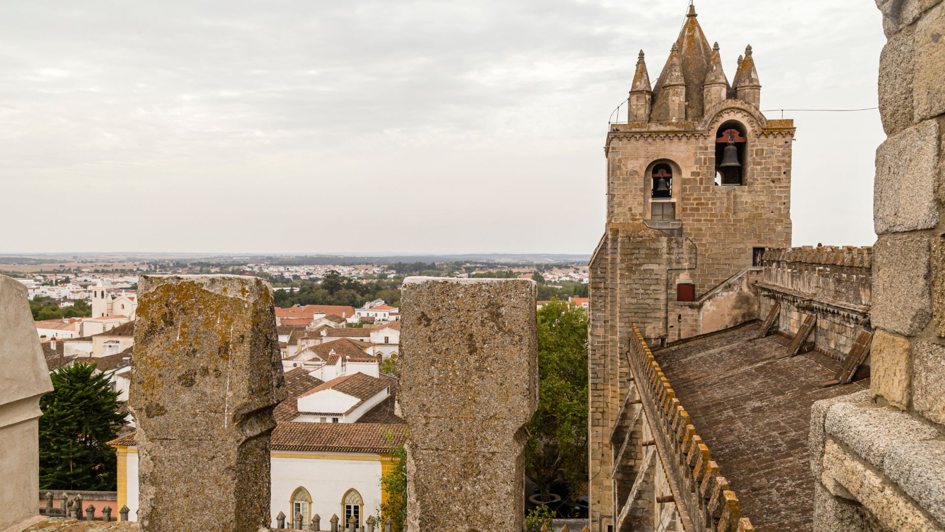 A high-angle view from the ramparts of the Cathedral of Évora in Portugal, showing its prominent bell tower and the tiled rooftops of the historic city below, under a cloudy sky.