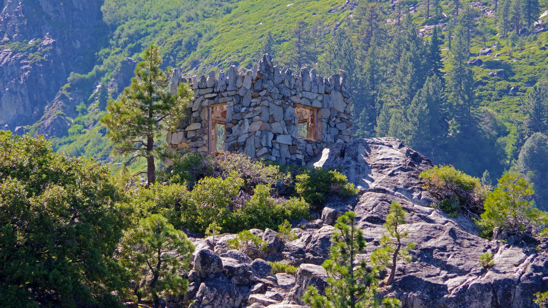 A stone tea house ruin with a crenellated roof, surrounded by trees and rocks, situated on a small island in a mountainous landscape.
