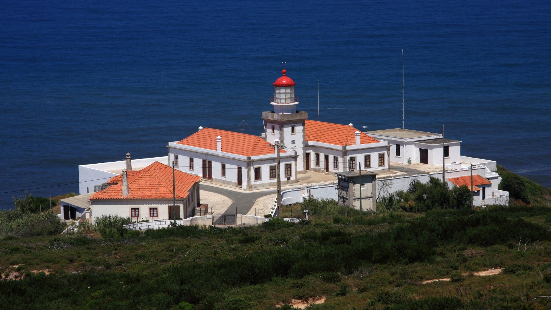 A high-angle view of the Farol do Cabo Mondego (Cape Mondego Lighthouse) complex in Portugal, featuring white buildings with red-tiled roofs and a red-domed lighthouse tower, situated on a green, brush-covered cliff overlooking the deep blue Atlantic Ocean.