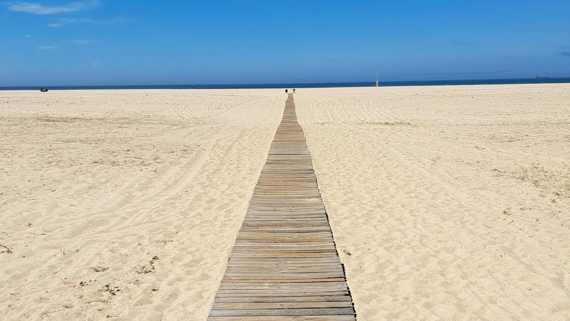 A long, straight wooden boardwalk extends across a wide, sandy beach towards the distant ocean under a clear blue sky. In the distance, a few small figures are visible on the boardwalk, and the horizon line where the sky meets the sea is distinct.