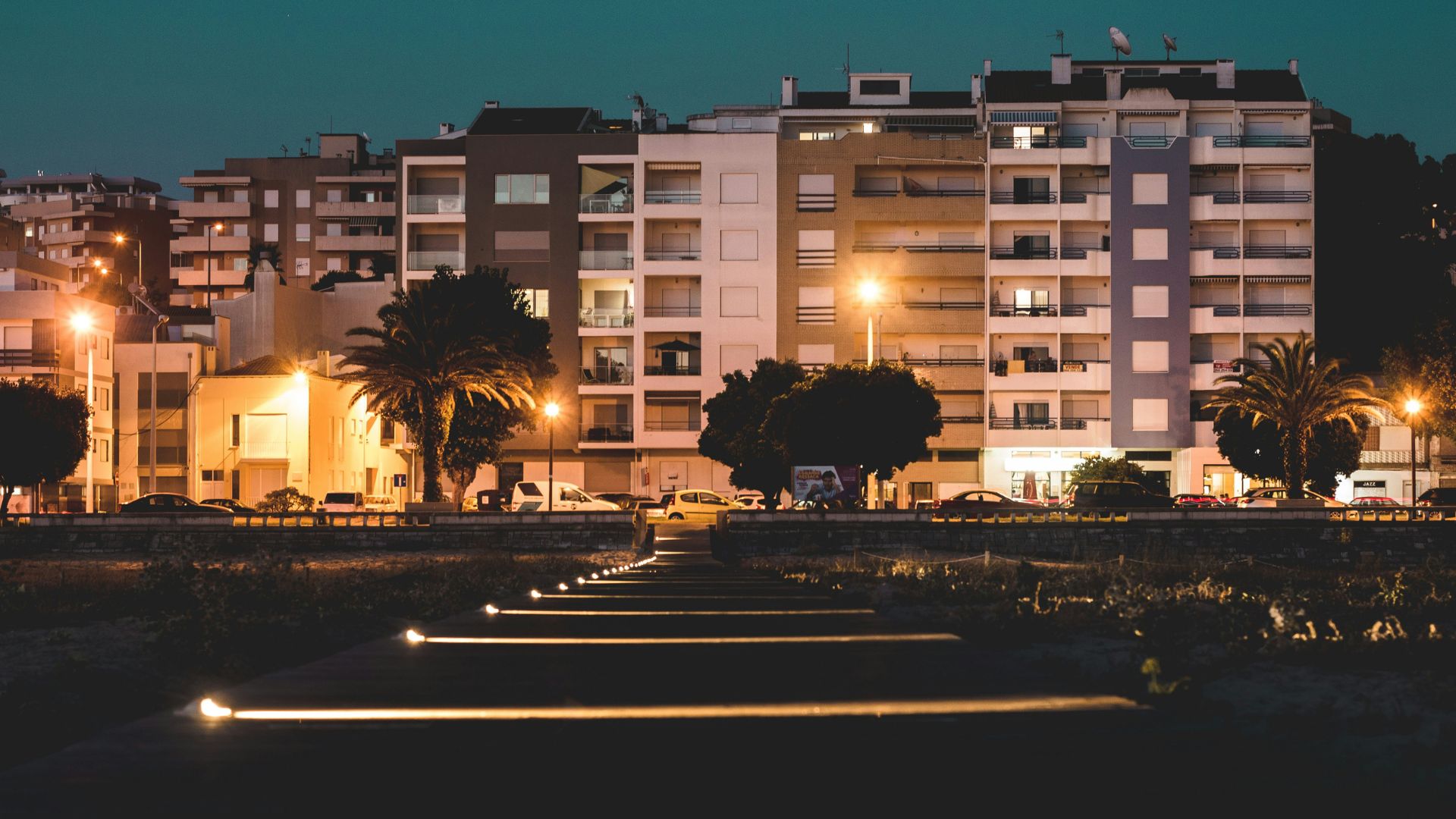 A nighttime view of illuminated buildings and a pathway with ground lights in a coastal town, featuring palm trees and streetlights, likely in Figueira da Foz, Portugal.