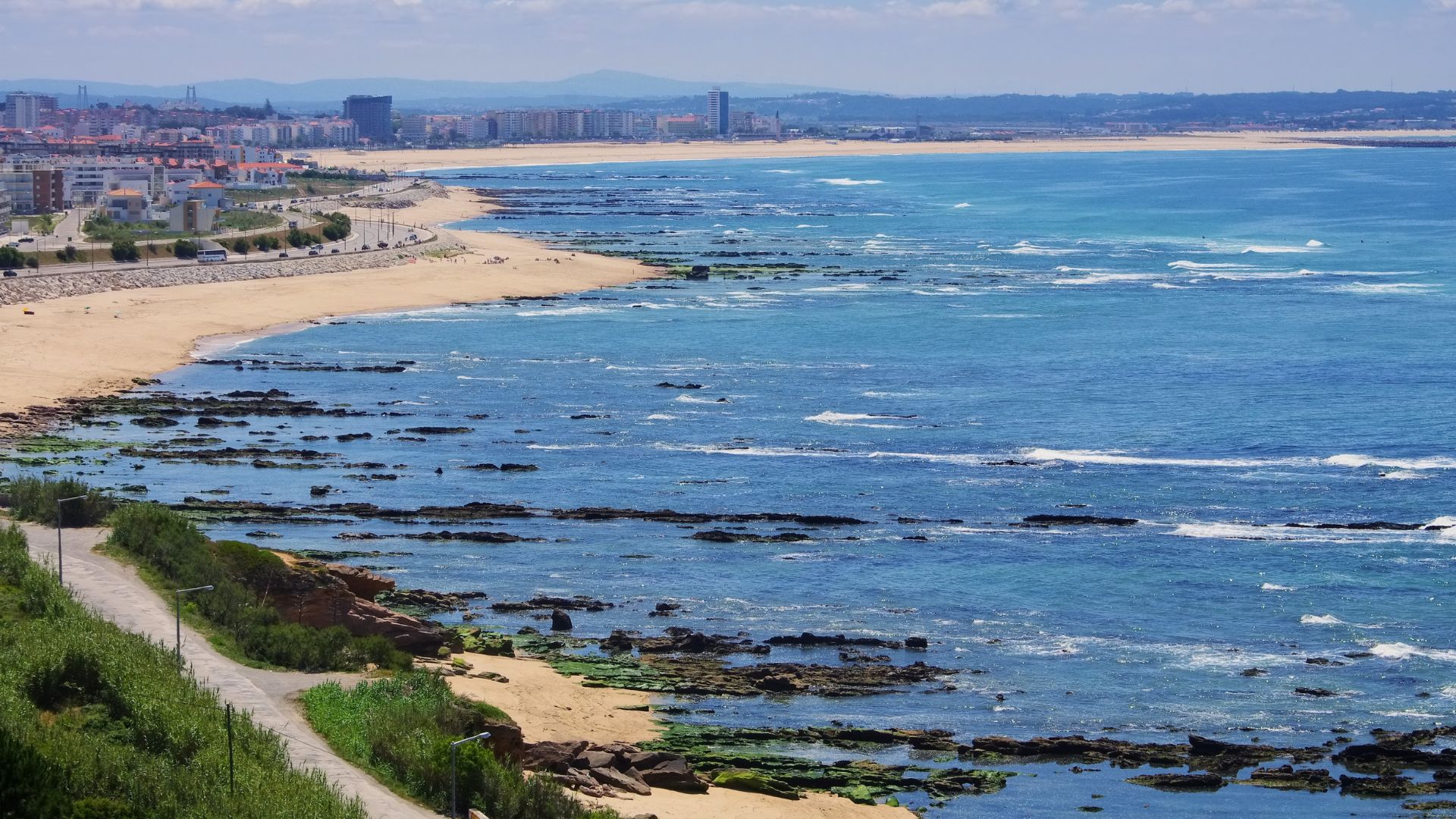 A panoramic view of the coastline of Figueira da Foz, Portugal, showing a sandy beach, a town with buildings along the shore, and a rocky intertidal zone extending into the blue Atlantic Ocean under a clear sky.