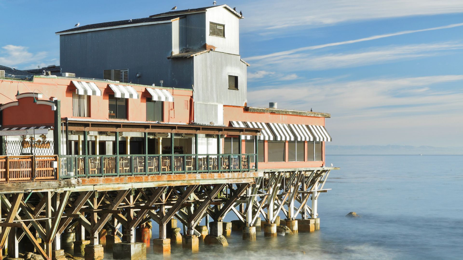 A waterfront building, identified as the Fish Hopper restaurant, stands on stilts over the ocean in Monterey, California, with a clear blue sky overhead and calm water below.