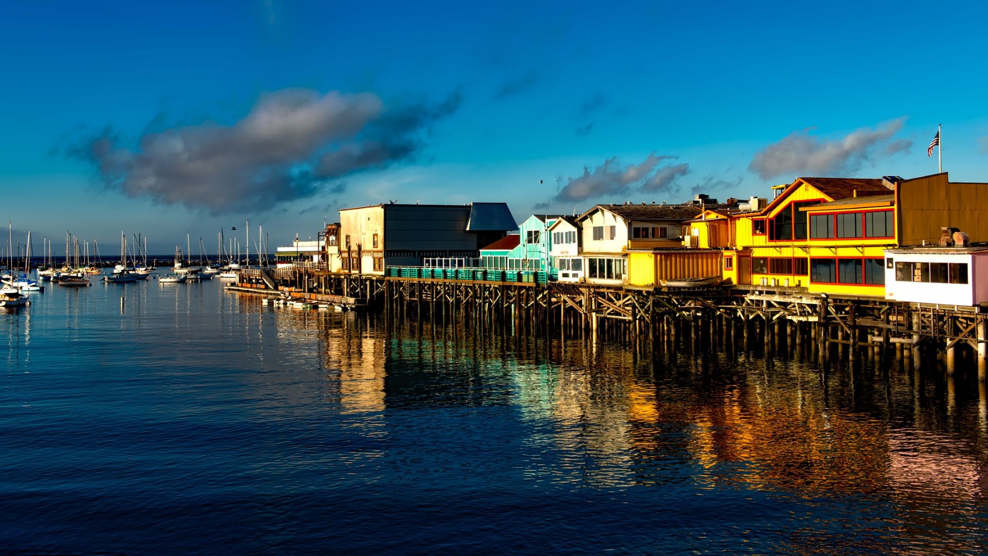 A vibrant image of Fisherman's Wharf in Monterey, California, featuring colorful buildings on stilts over calm blue water under a bright sky with scattered clouds, with boats docked to the left.