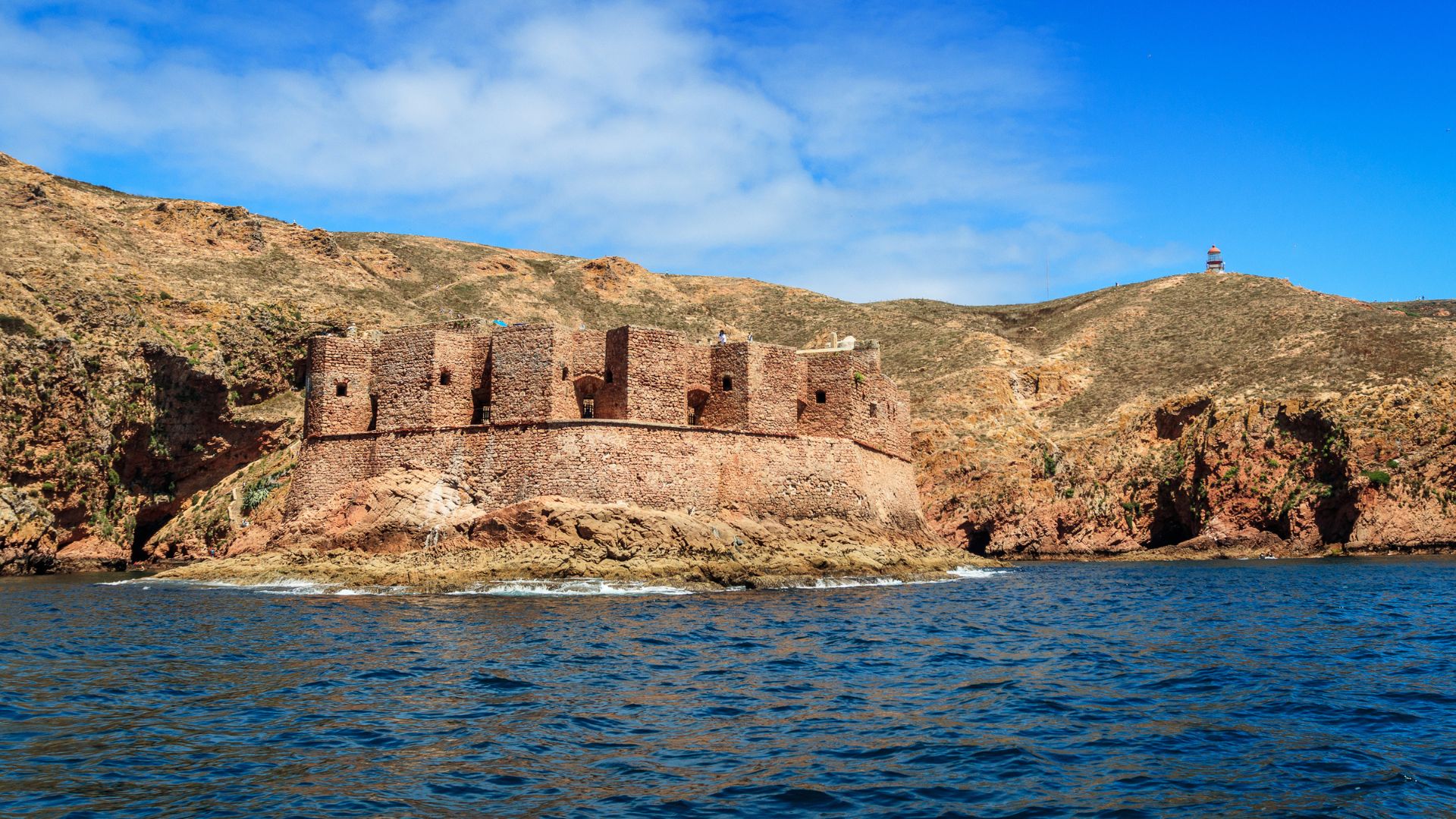A historic stone fortress, the Fort of São João Baptista, stands on a small rocky island in the middle of clear blue water, with rugged, hilly terrain rising behind it under a bright blue sky.
