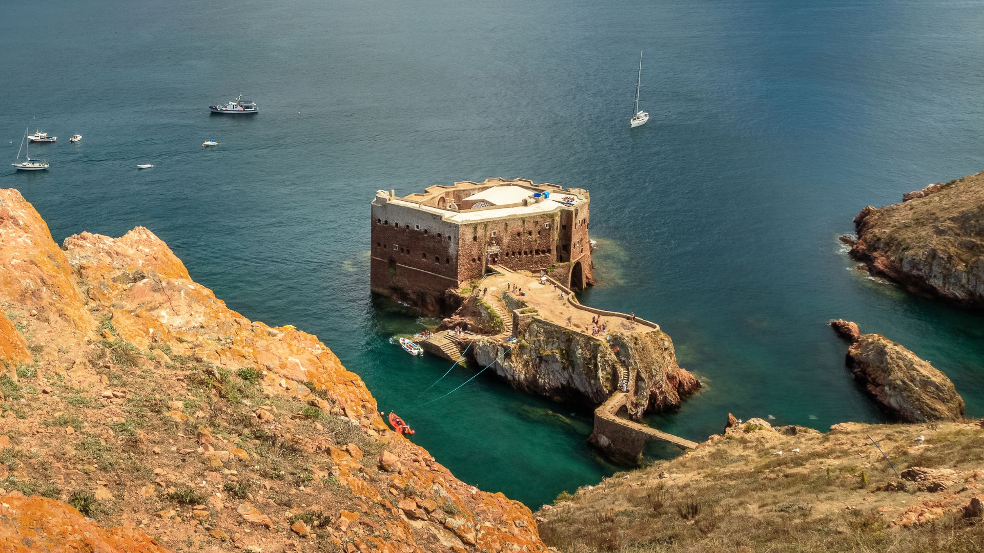 A high-angle view of the historic Fort of São João Baptista, a stone fortress built on a small rocky islet connected by a narrow causeway to the larger Berlenga Grande island, surrounded by the clear blue waters of the Atlantic Ocean under a bright sky.