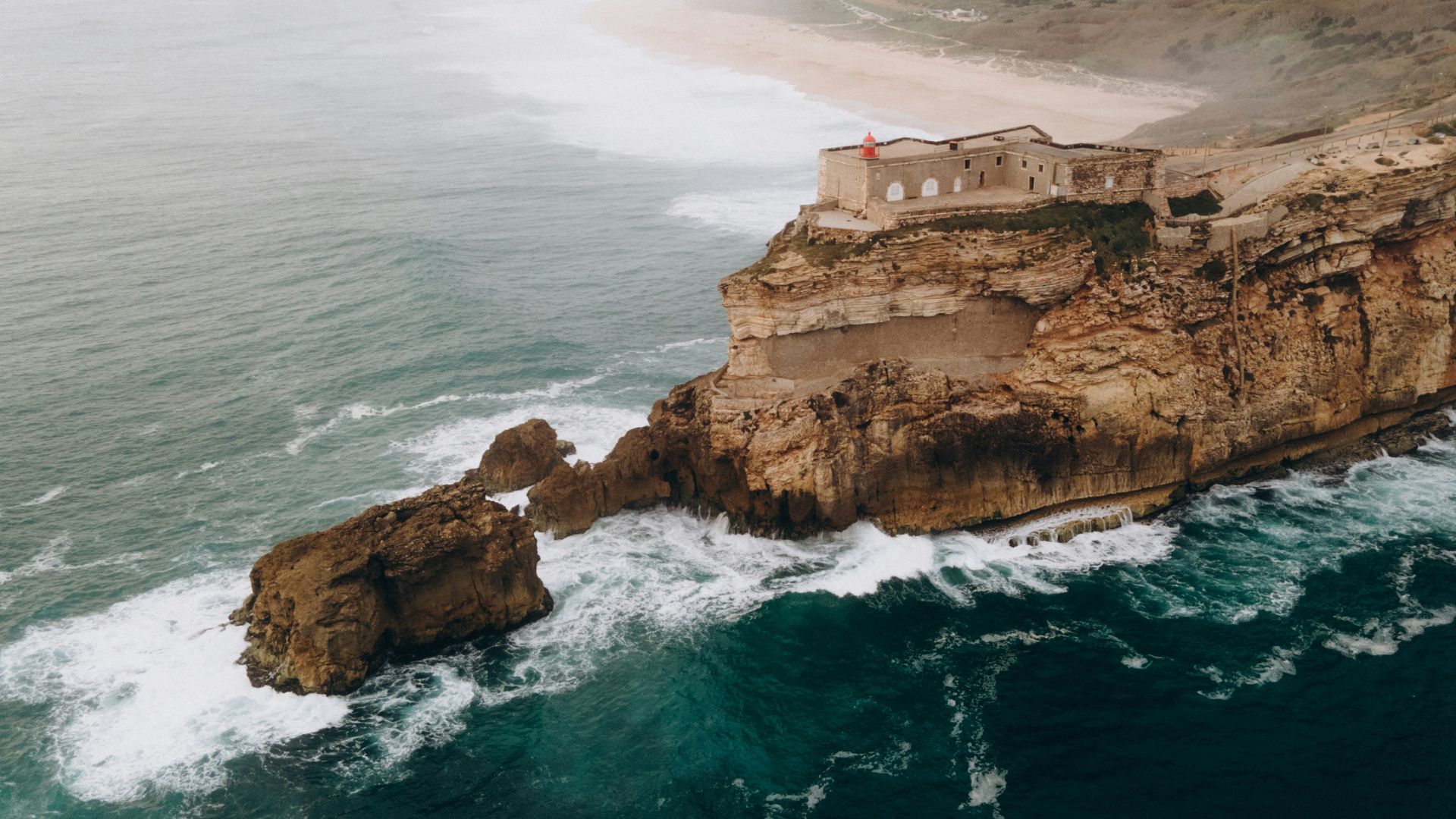 An aerial view of the Forte de São Miguel Arcanjo and its lighthouse perched on a rugged cliff overlooking the turbulent, dark blue waters of the Atlantic Ocean and the expansive, sandy stretch of Praia do Norte beach in Nazaré, Portugal.