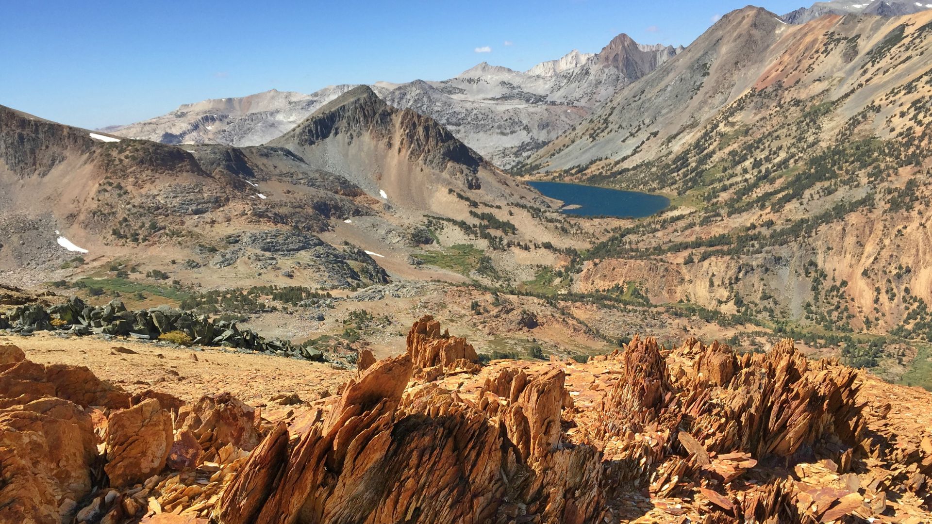 A wide-angle landscape shot captures a high-altitude mountain scene dominated by rugged, rocky terrain in the foreground, featuring reddish-brown rock formations. In the middle ground, a deep blue alpine lake is nestled within a valley surrounded by towering, multi-colored peaks, some still holding patches of snow. The distant mountains are hazy, suggesting vastness and elevation. The sky is clear and blue.
