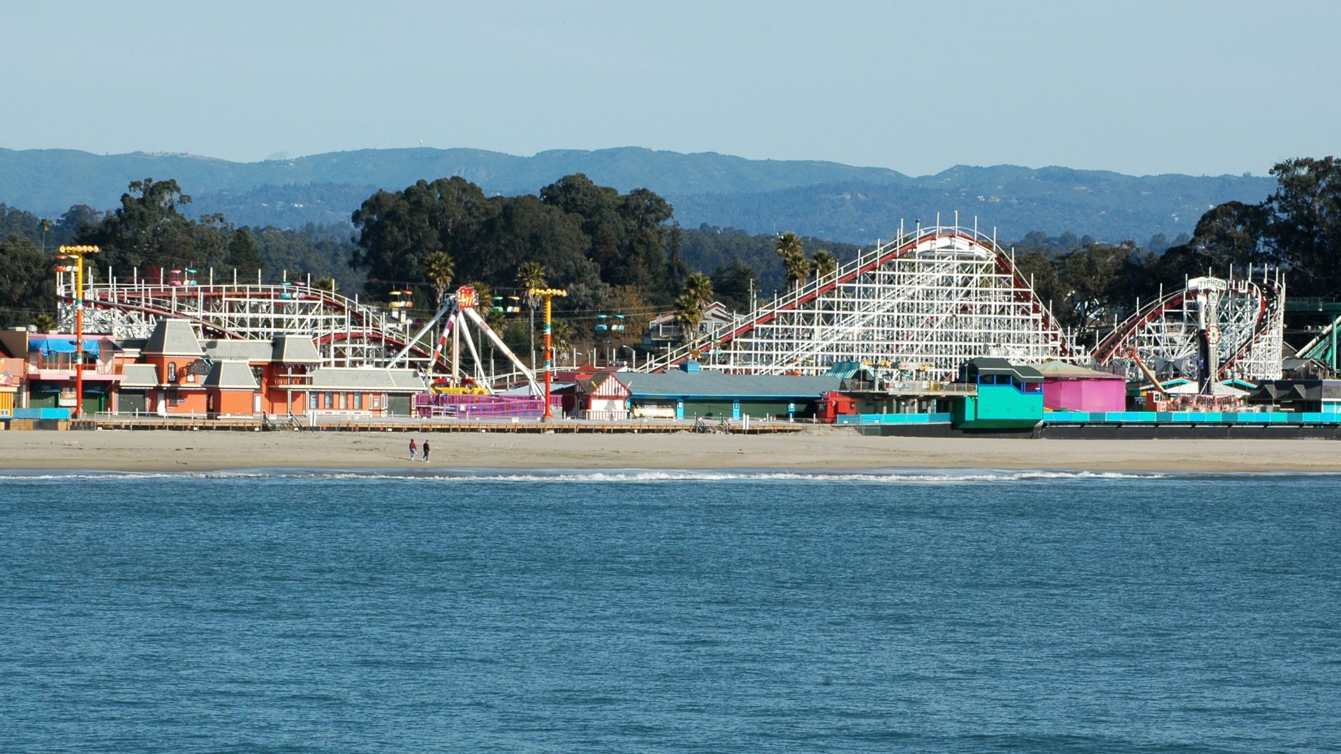 A vibrant image of the Santa Cruz Beach Boardwalk, showcasing the classic wooden roller coaster, the Giant Dipper, alongside other amusement park rides and buildings, all situated on a sandy beach next to the ocean, with mountains visible in the background under a clear sky.