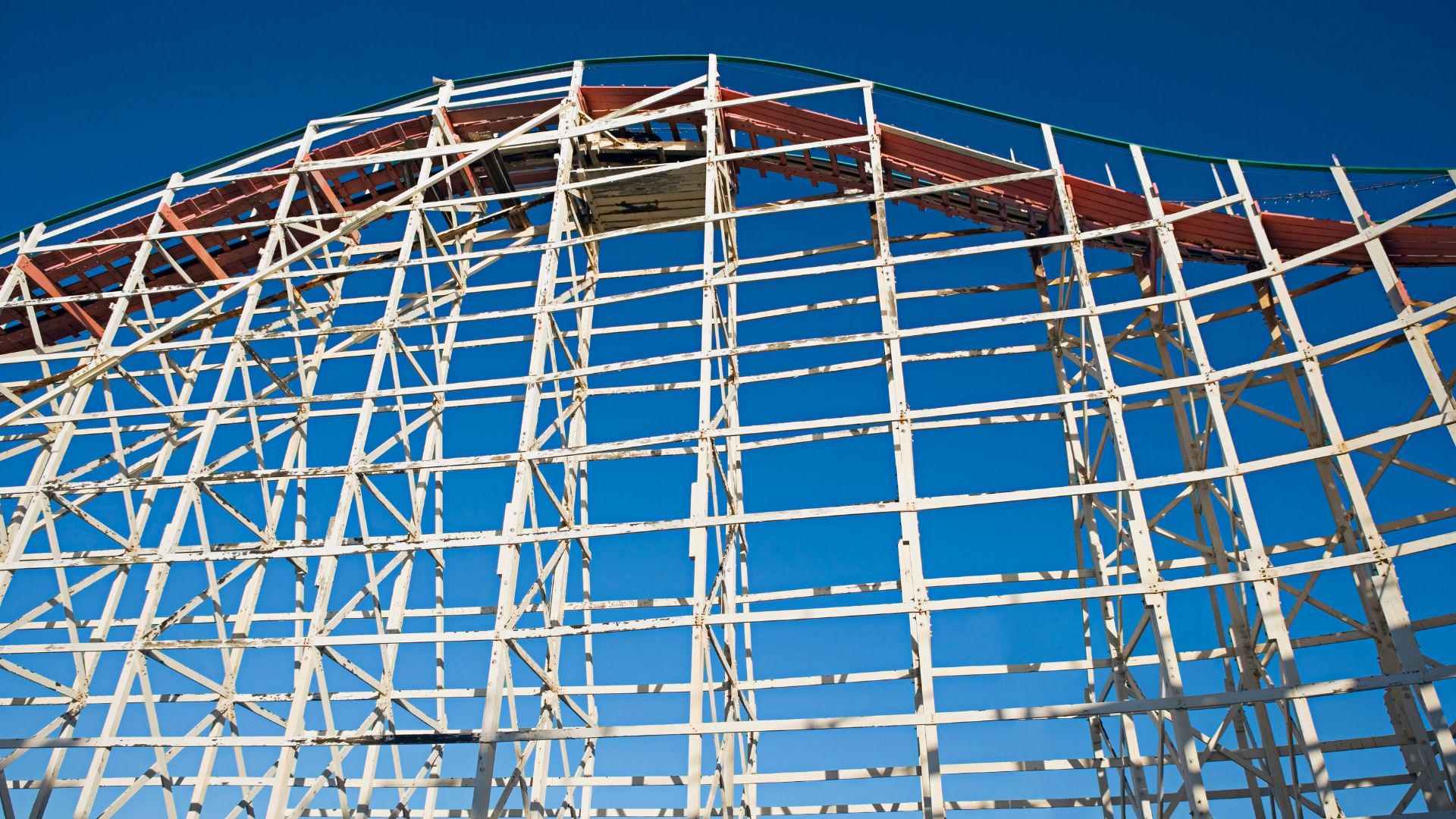 A close-up view of the white wooden structure of a roller coaster against a clear blue sky, with parts of the reddish-brown track visible at the top. 