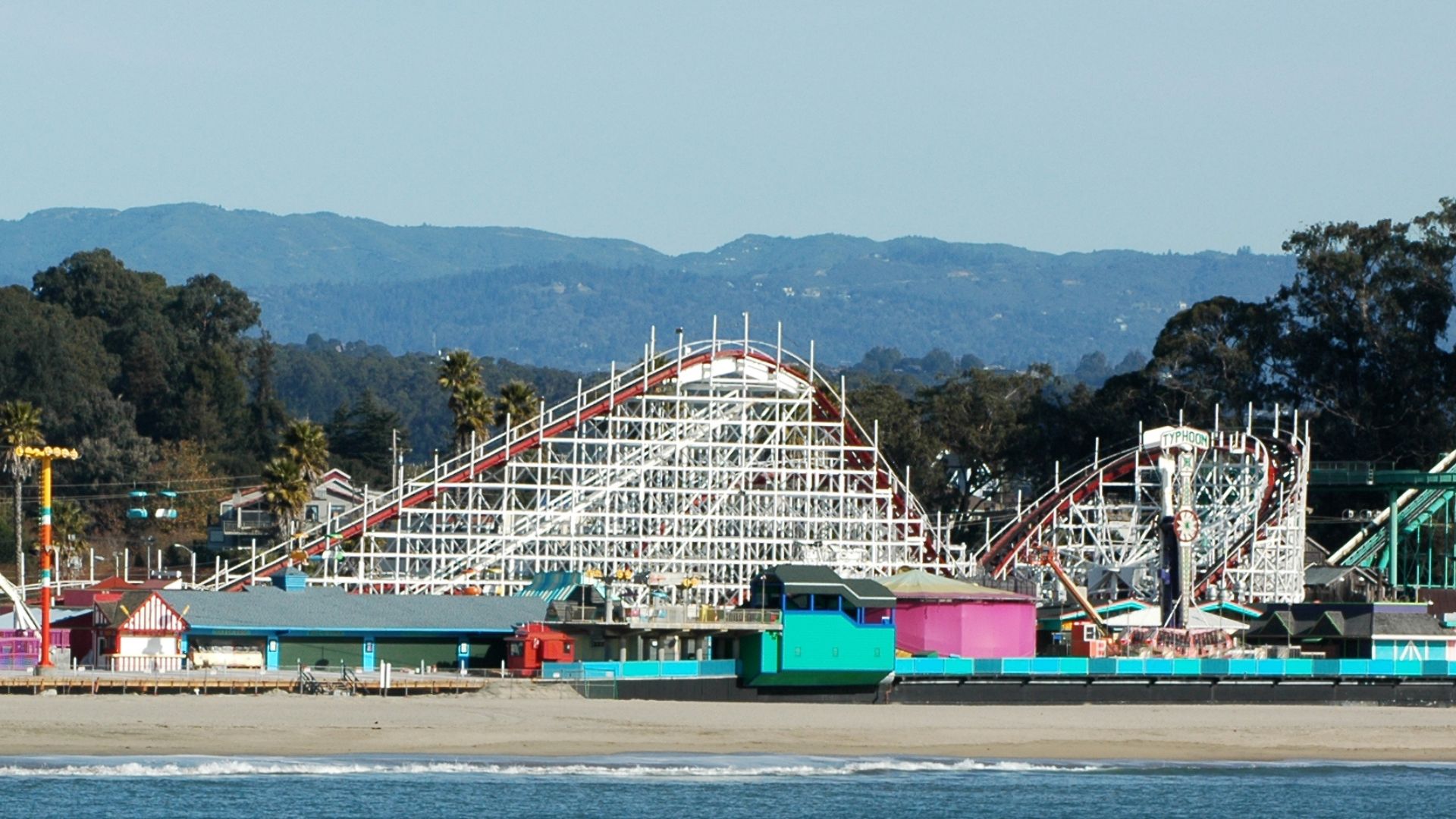 A classic wooden roller coaster with white supports and red tracks, known as the Giant Dipper, stands prominently on a sandy beach alongside colorful buildings, with mountains visible in the background under a clear sky.