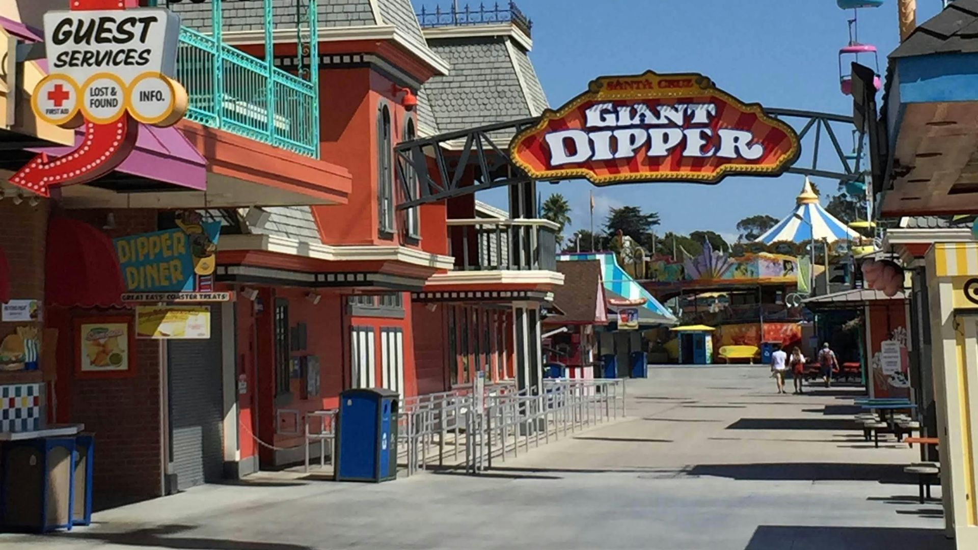 A vibrant outdoor scene at the Santa Cruz Beach Boardwalk under a clear blue sky, showing the entrance to the Giant Dipper roller coaster, a Guest Services building with a prominent ice cream cone sculpture, and colorful sky ride gondolas overhead.night