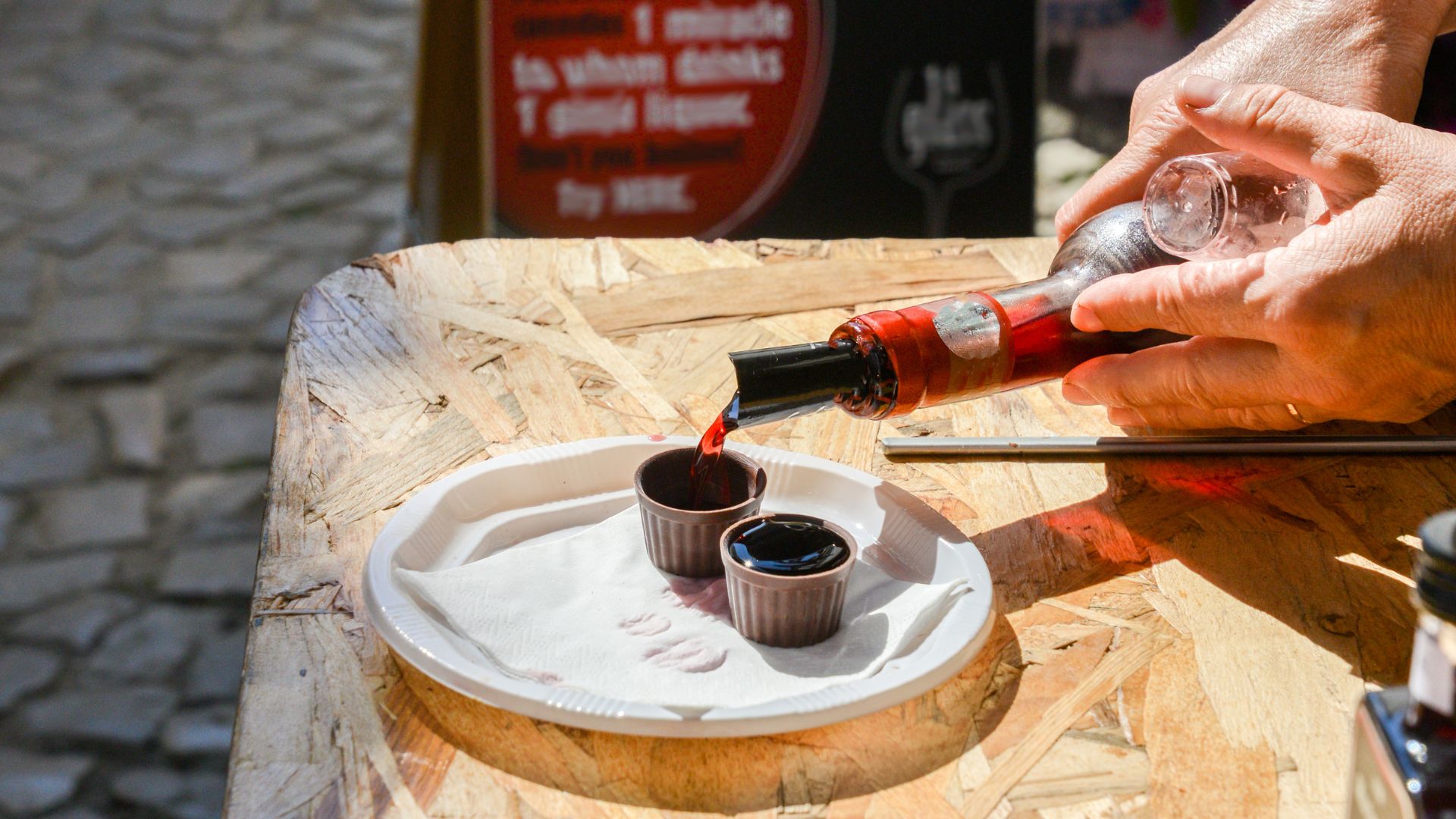 A close-up shot of hands pouring dark red Ginja de Óbidos cherry liqueur from a bottle into small, edible chocolate cups on a white plate, placed on a rustic wooden table.