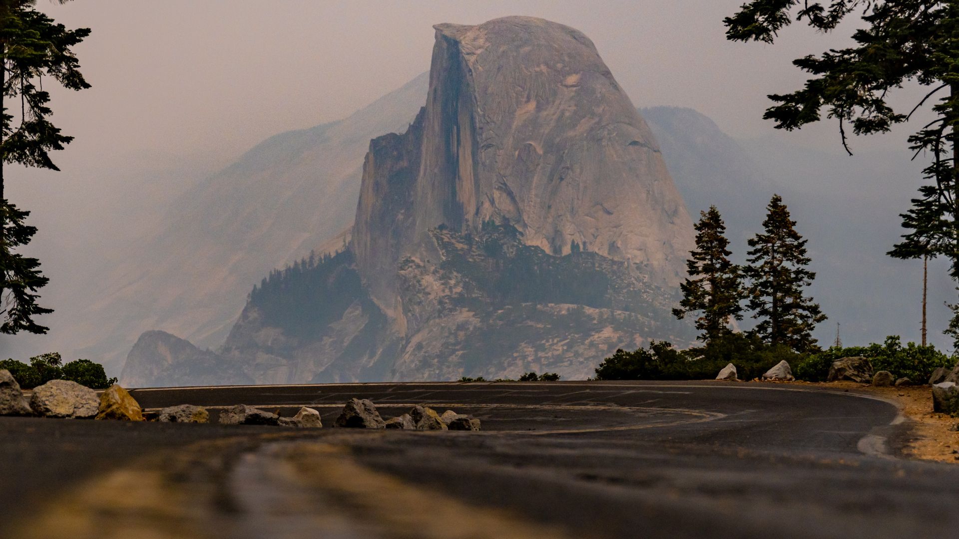 A wide shot showing Half Dome, a prominent granite formation, in the background, viewed from a paved overlook (likely Glacier Point) with a road and scattered rocks in the foreground, all under a hazy sky.