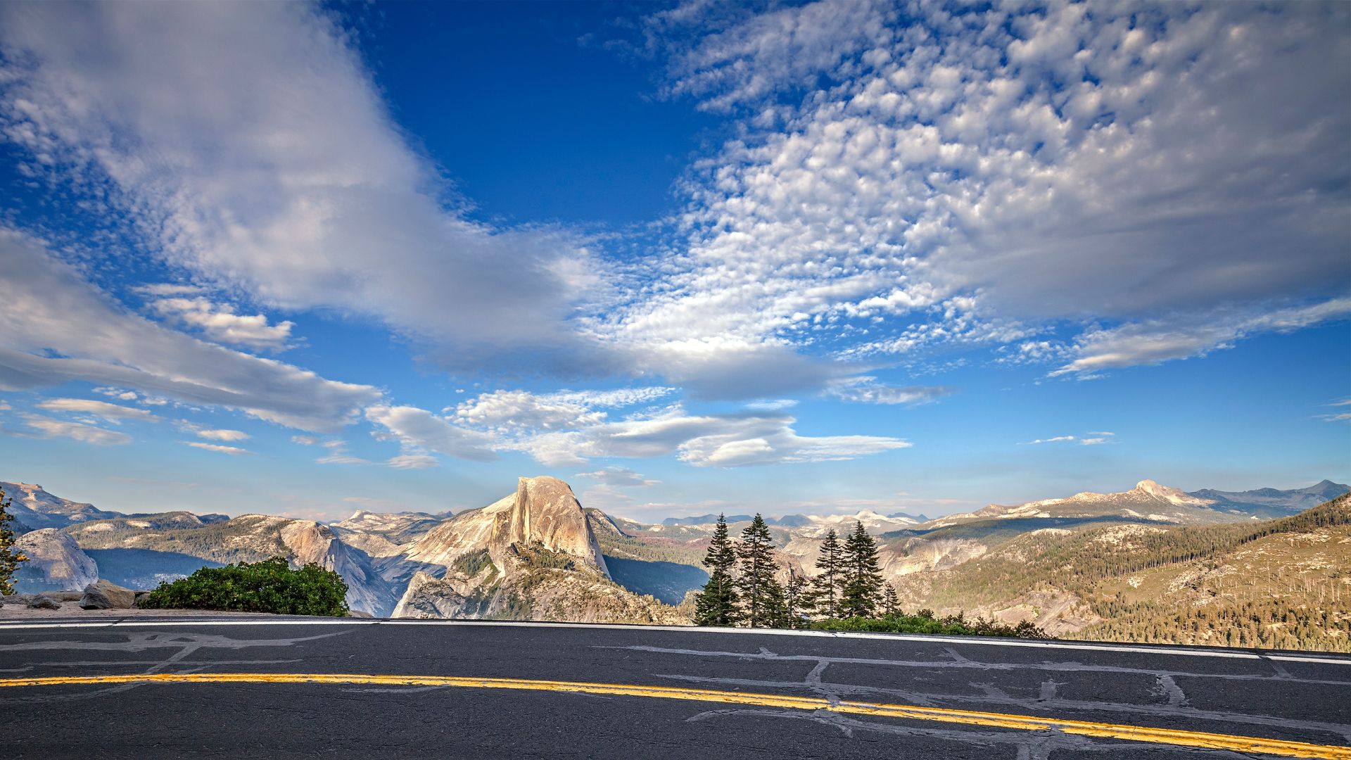 A scenic view from Glacier Point Road in Yosemite National Park, showing the iconic Half Dome mountain peak under a blue sky with scattered clouds.
