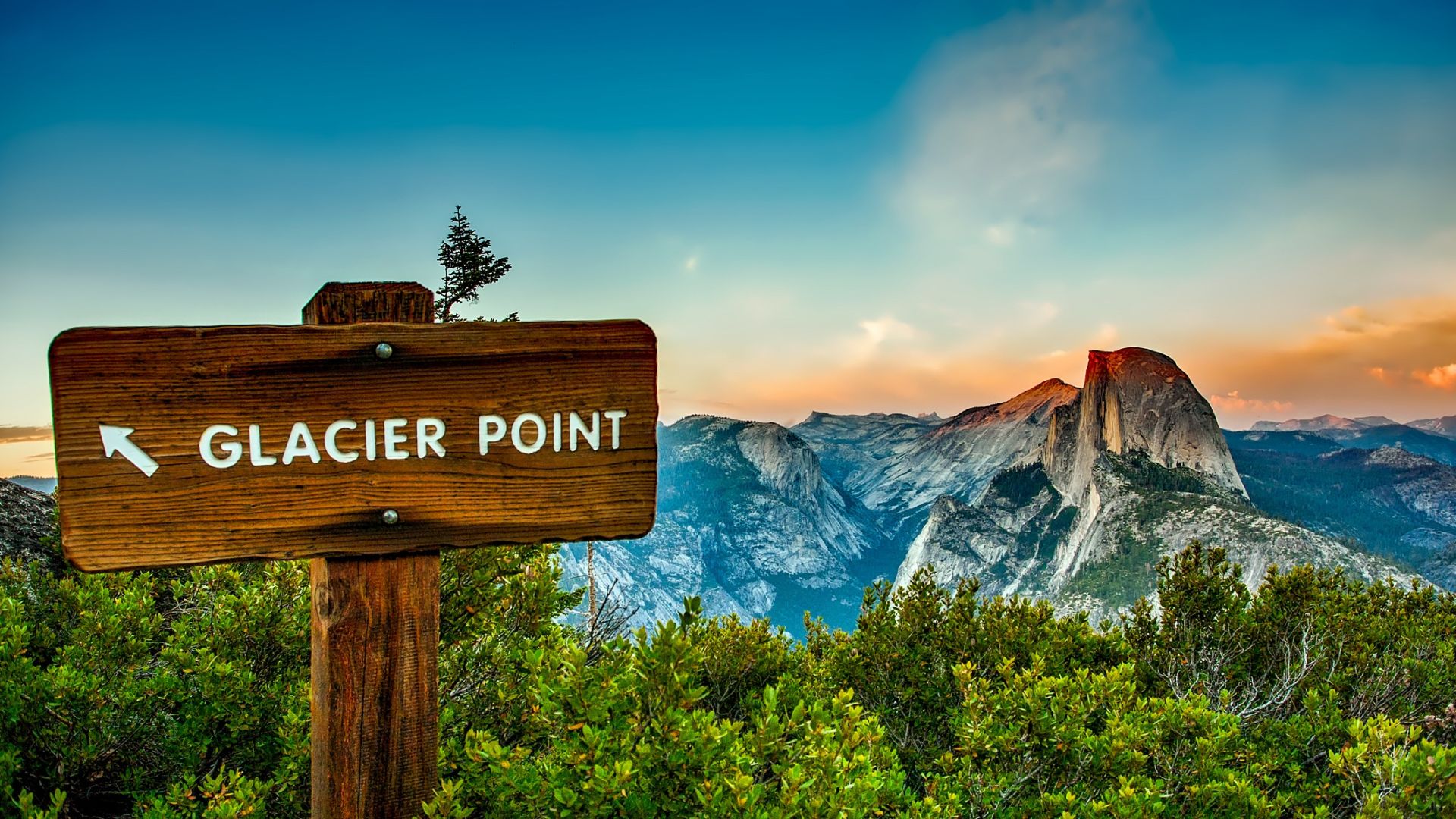 A wooden sign pointing left to "GLACIER POINT" stands in the foreground, with the iconic Half Dome and other mountainous terrain of Yosemite National Park visible in the background under a dramatic sky. 