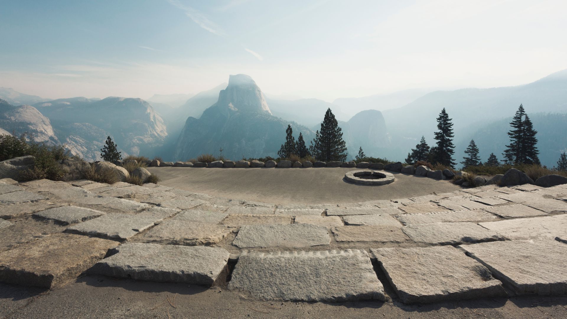 A wide-angle view of Glacier Point, featuring a stone amphitheater in the foreground with a fire pit, overlooking a vast mountain landscape with prominent peaks and hazy skies in the distance, including Half Dome.