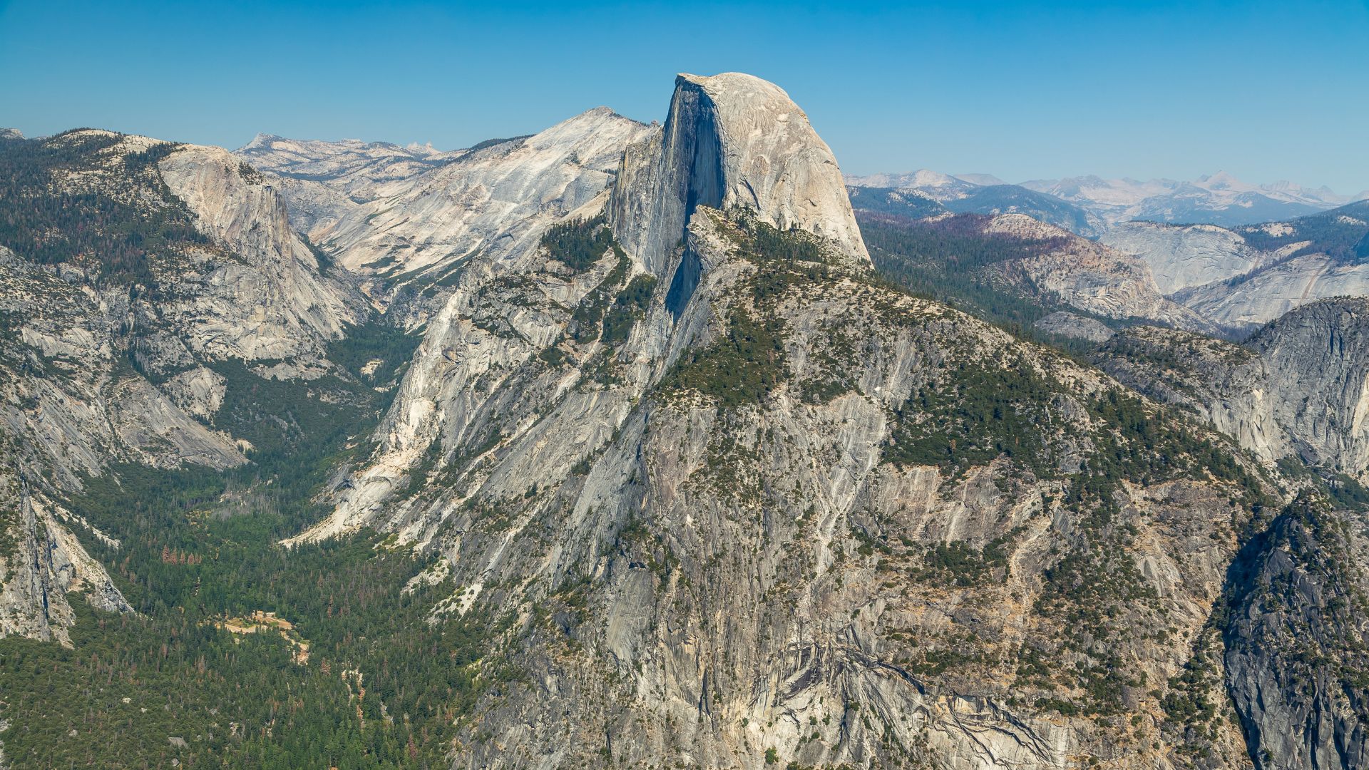 An aerial view of Half Dome, a distinct granite rock formation with a sheer vertical face, rising above a forested valley in Yosemite National Park, California, under a clear blue sky. The surrounding landscape features other rugged, tree-covered mountains and valleys.