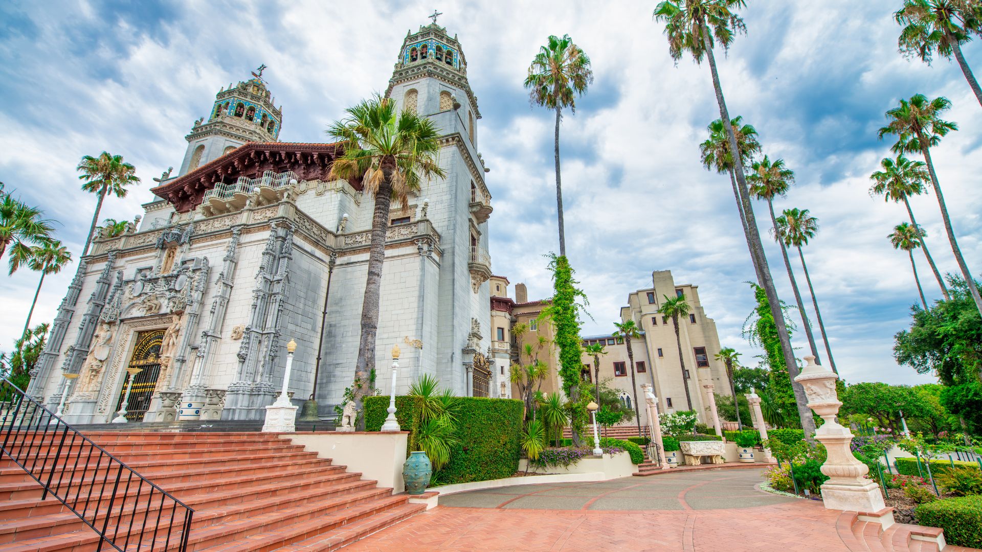 A wide, low-angle shot captures the grandeur of Hearst Castle, a sprawling, ornate white building with multiple towers and intricate architectural details, set against a partly cloudy sky. In the foreground, a wide, terraced staircase leads up to the main entrance, flanked by lush green gardens and tall, slender palm trees that frame the castle and extend into the background. 